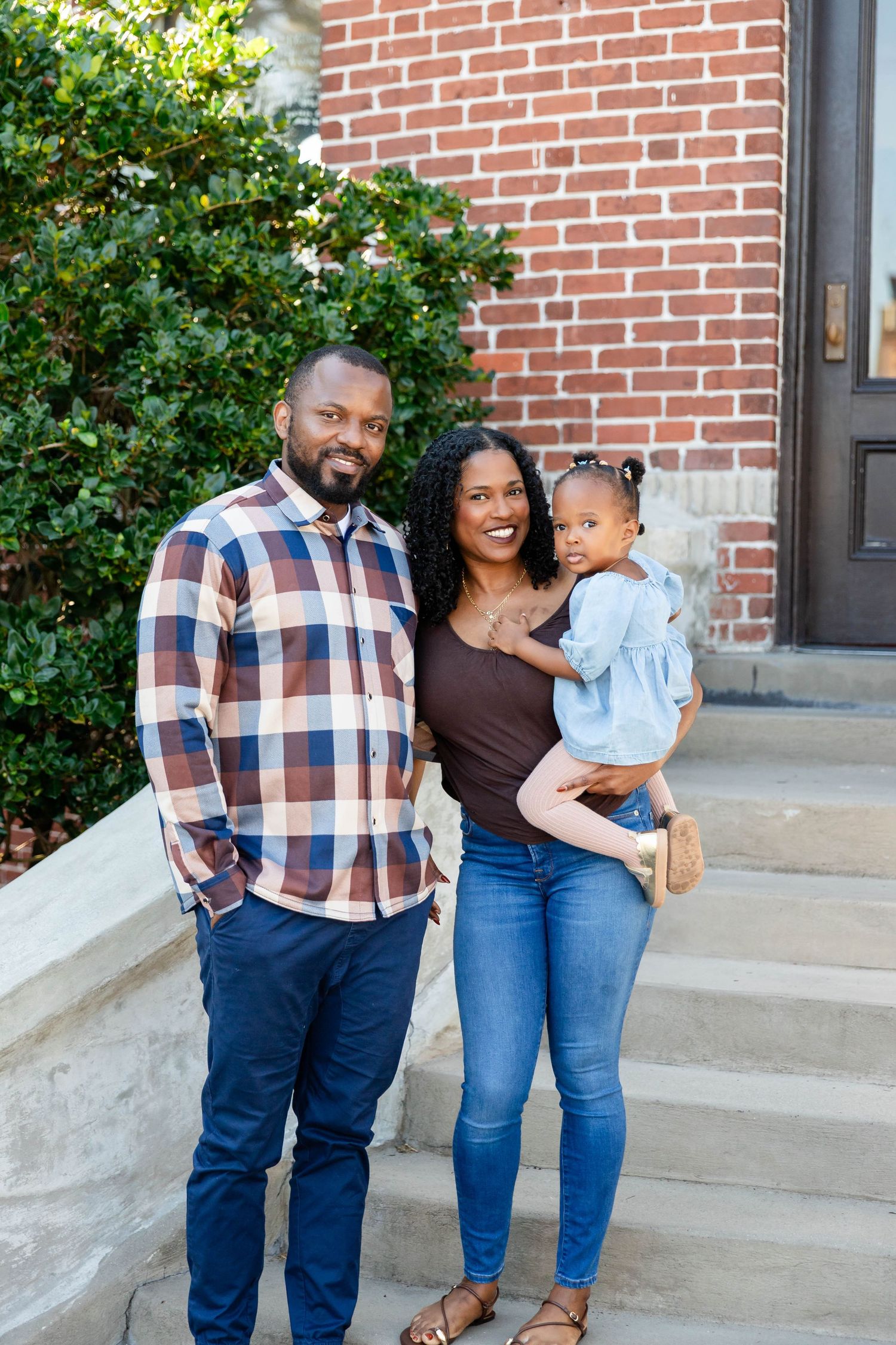 Black family of three poses on brick building steps, parents smiling with toddler girl in blue dress at the University of Tampa.