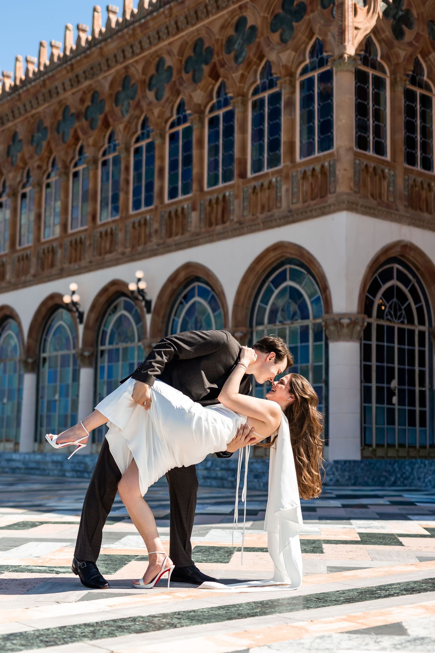 Groom dips bride for a kiss outside ornate Venetian-style building with arched windows on wedding day at The Ringling Museum.