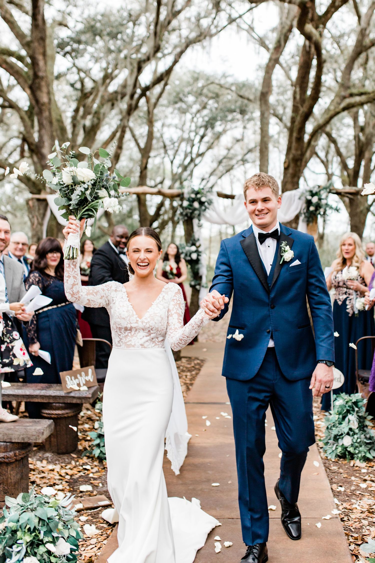 Joyful bride raises bouquet as she and groom walk back down the aisle after their outdoor woodland ceremony.