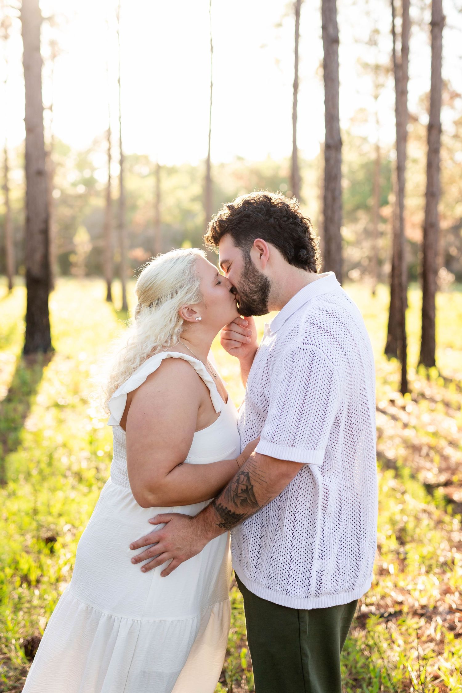 Couple sharing a romantic kiss in a sunlit forest, woman in white dress, man in white shirt with tattoos.