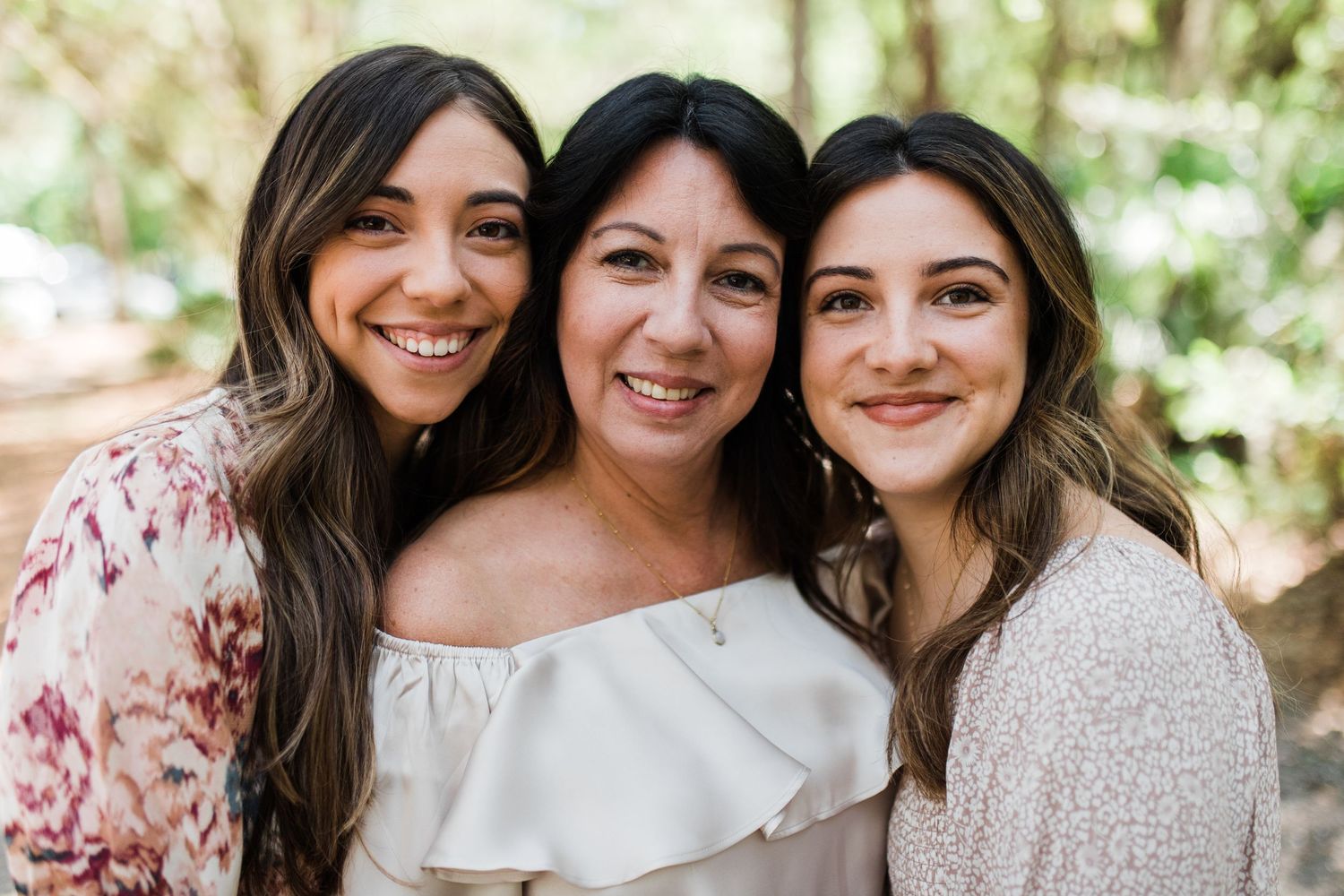 Three smiling women pose together outdoors in a warm family portrait among green trees.