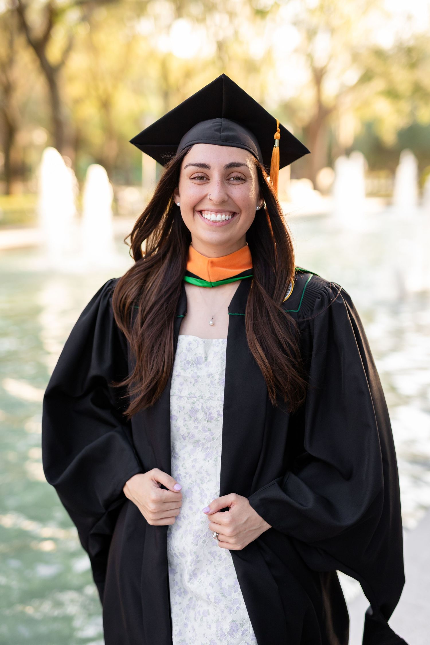 Smiling female graduate in black cap and gown with orange and green hood standing near a fountain at the University of South Florida.