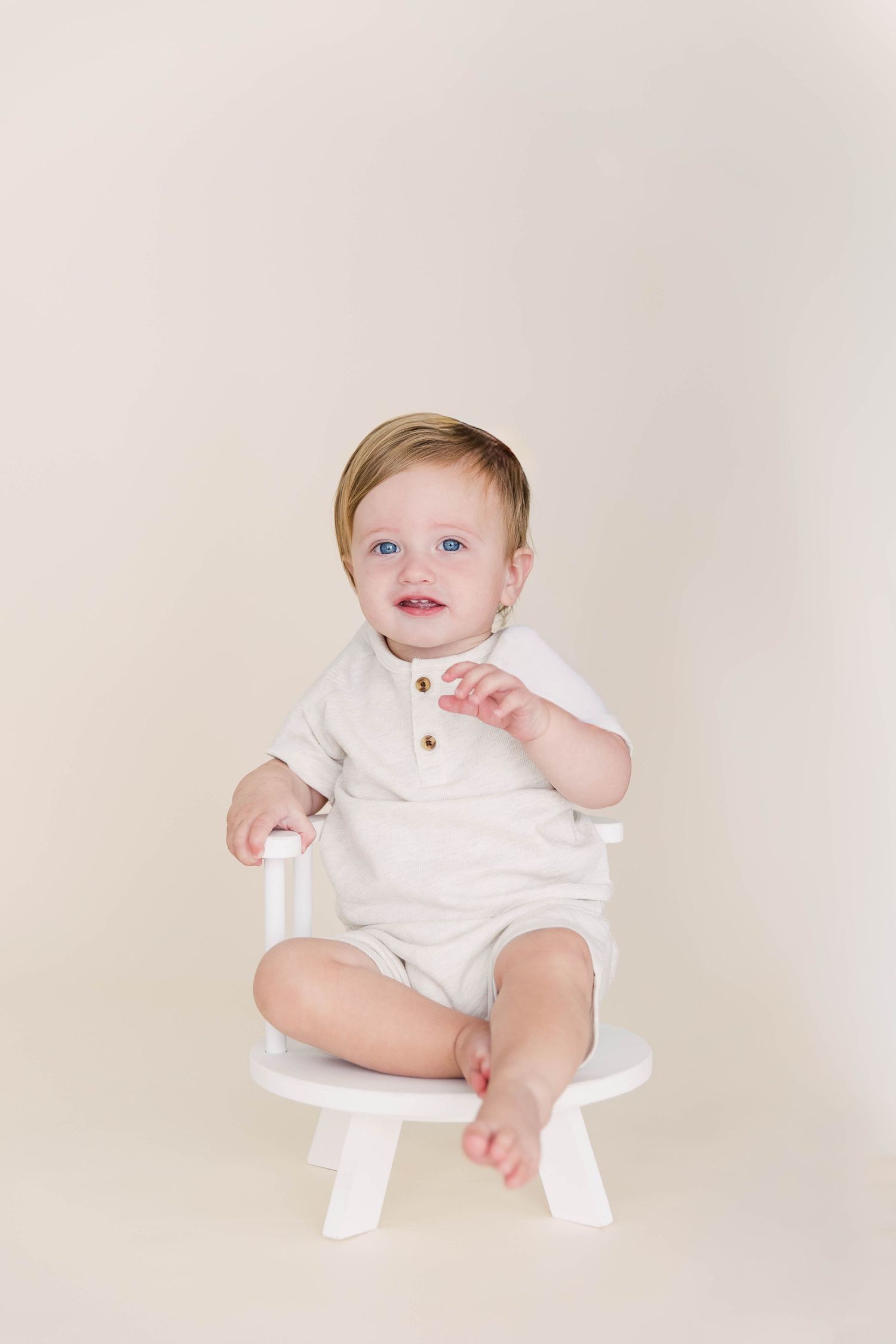 An adorable blue-eyed baby in a cream romper sits on a small white chair.