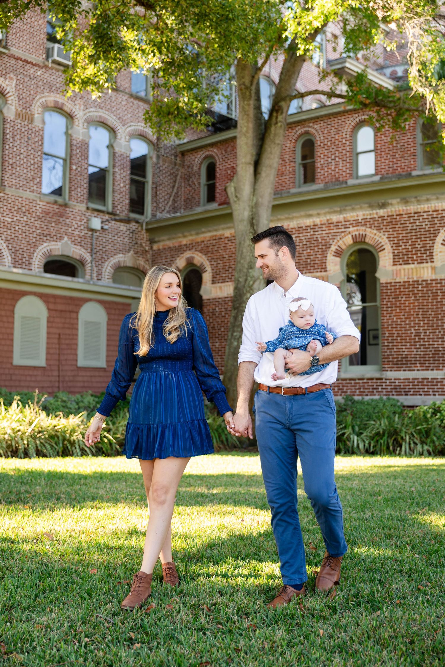 Happy couple walks holding hands with baby in blue outfit on sunny lawn by brick building at the University of Tampa..