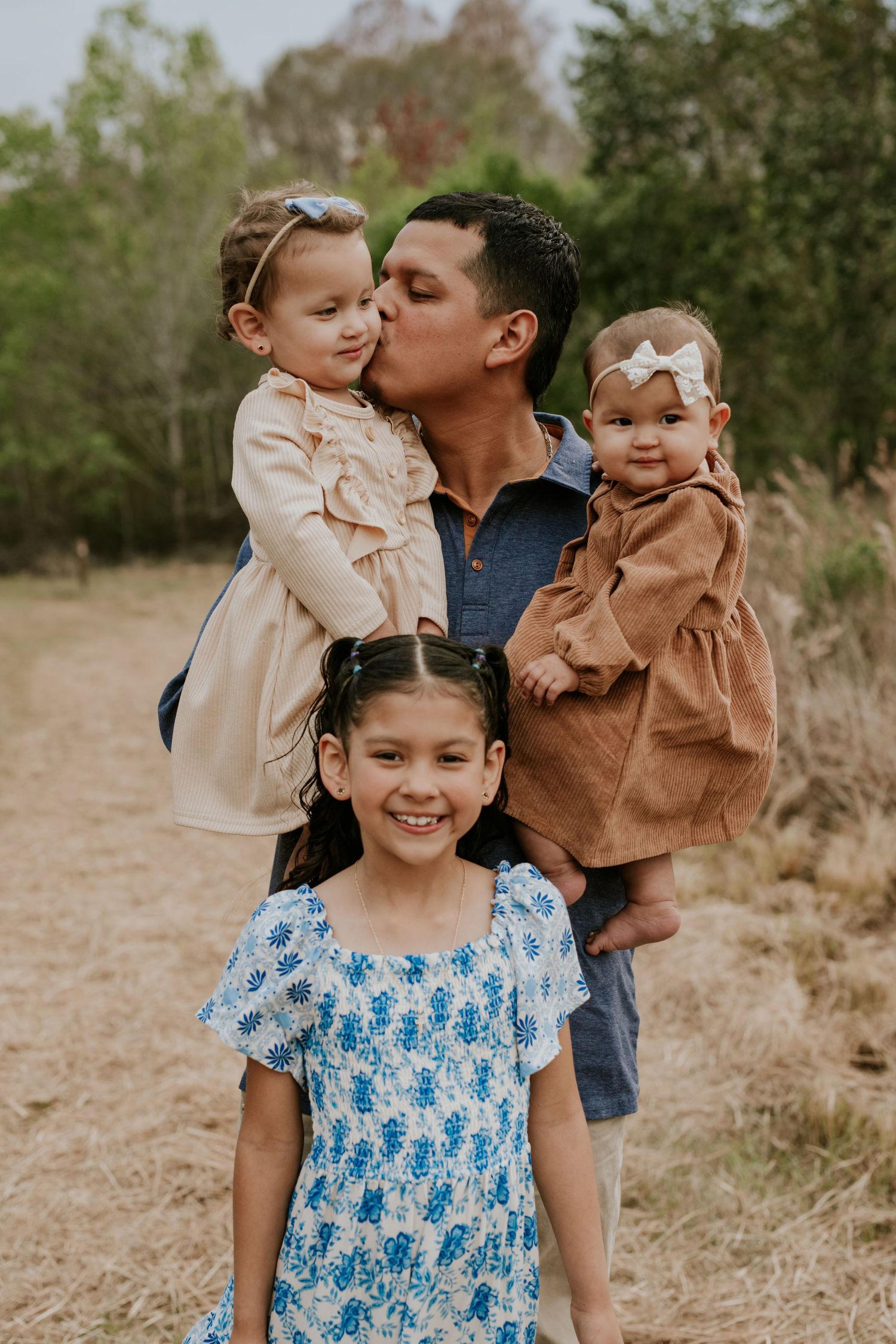 Loving father kisses toddler girl while holding baby and older daughter poses in front.