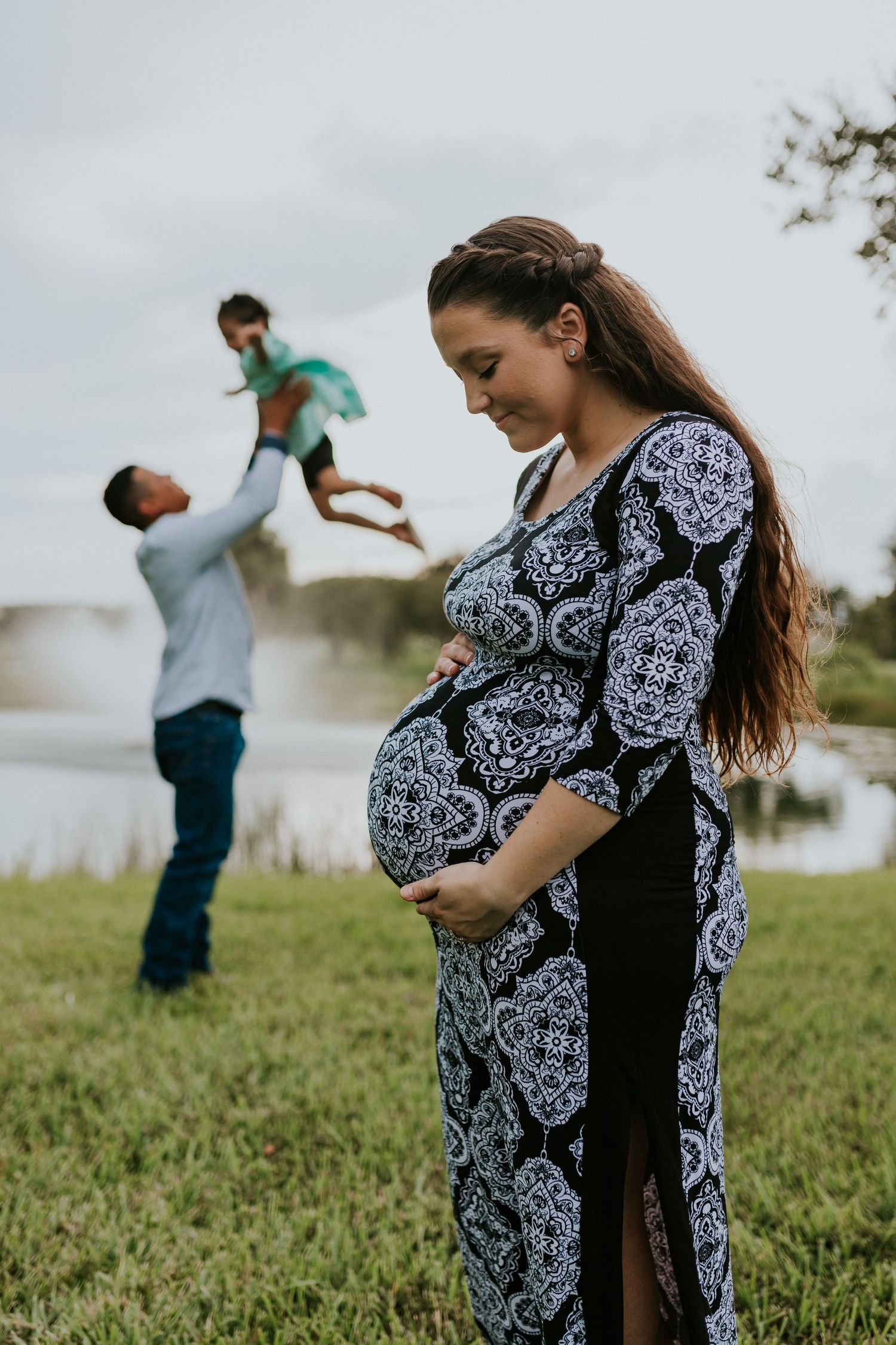 A pregnant woman in a black and white dress cradles her belly while her partner plays with their child outdoors.