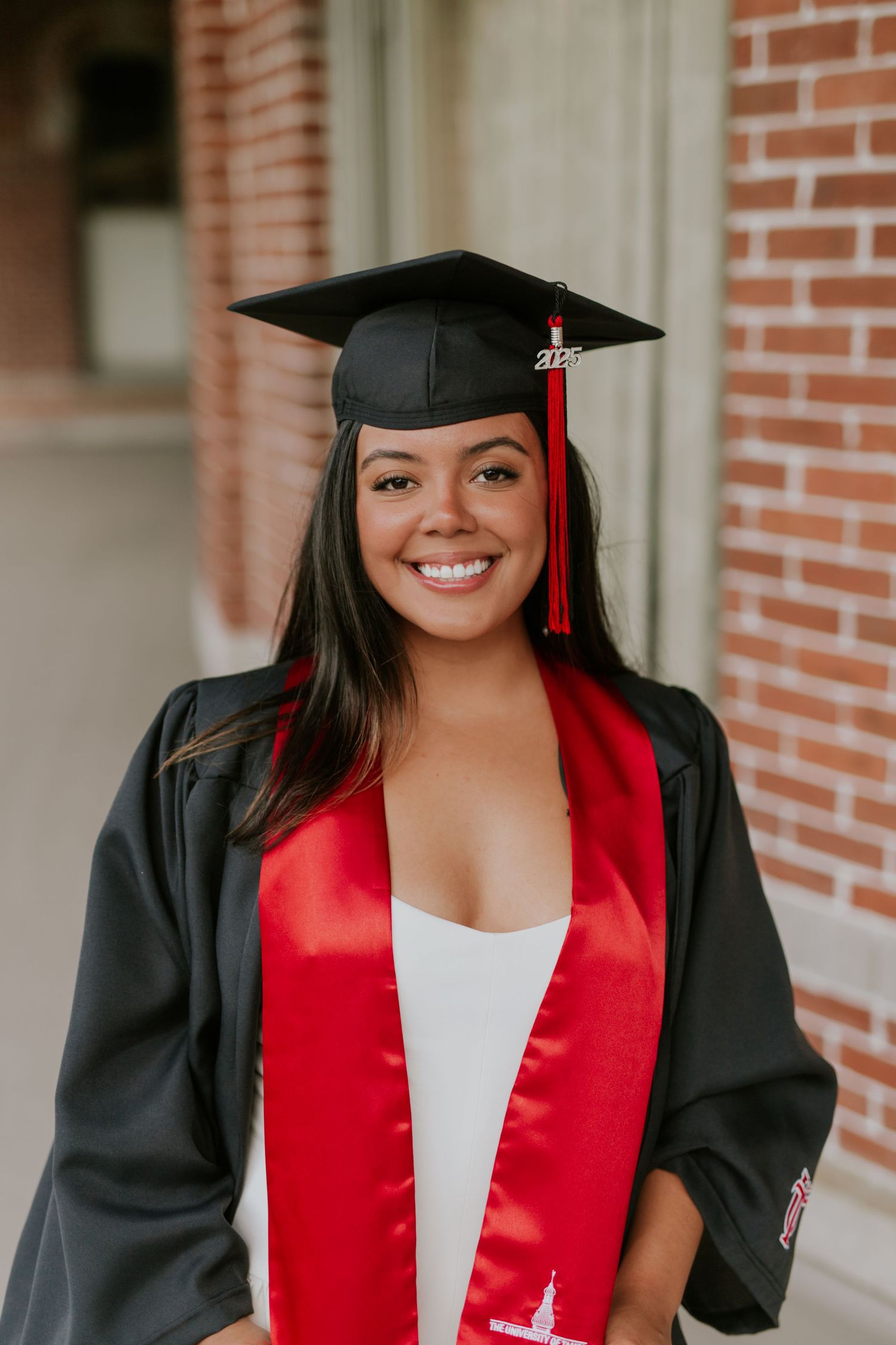 Smiling young woman in black graduation cap and gown with red sash poses outdoors near brick building.