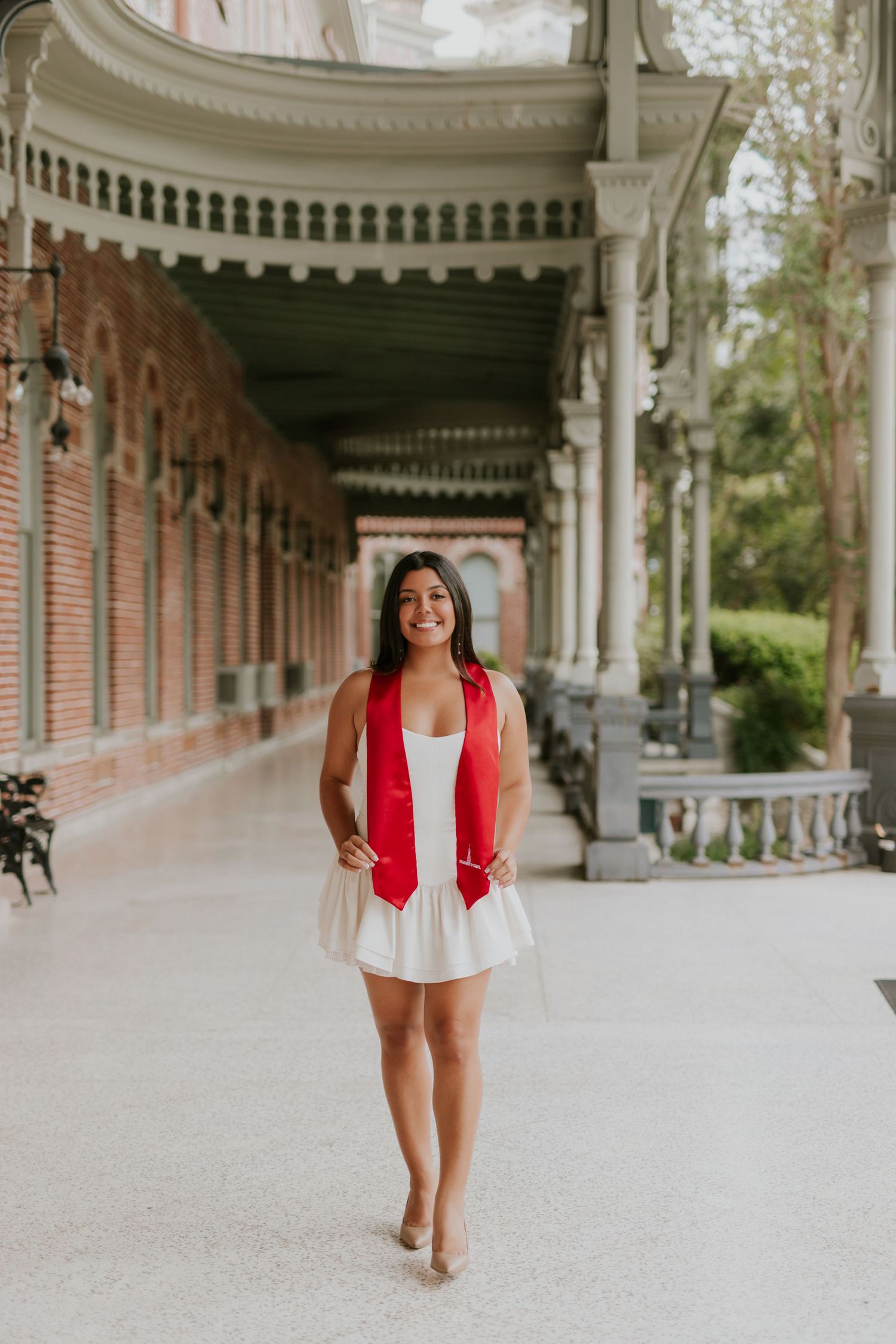 Young woman in white dress and red graduation stole smiling in ornate brick colonnade walkway.