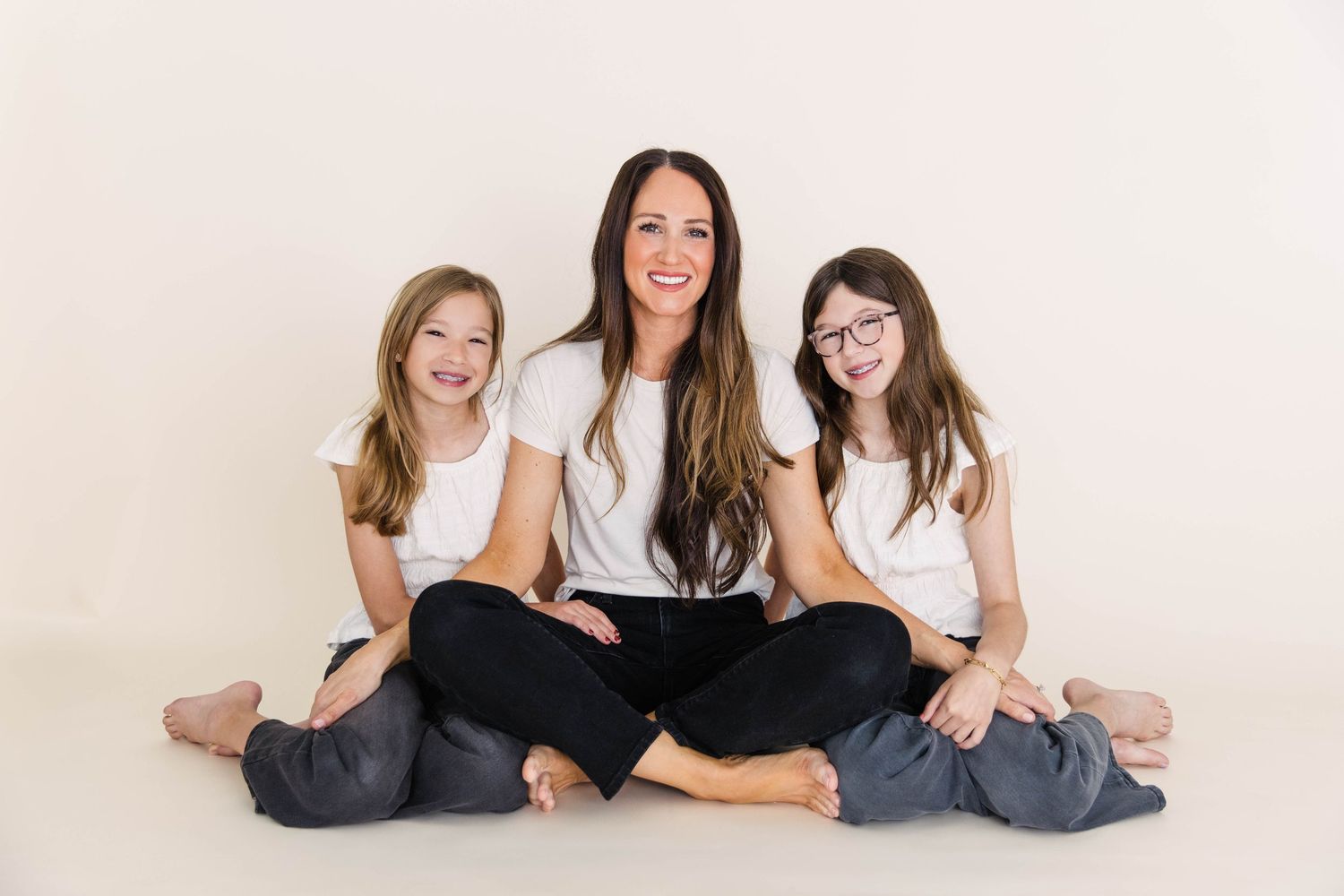 Brunette mom smiling with two daughters in white tops and black pants on studio floor.