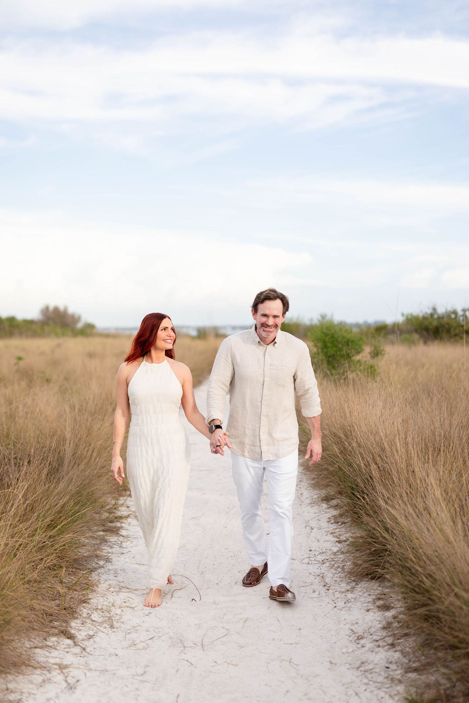 Smiling couple walking hand in hand along a sandy path through tall golden grass under a cloudy sky at Honeymoon Island.