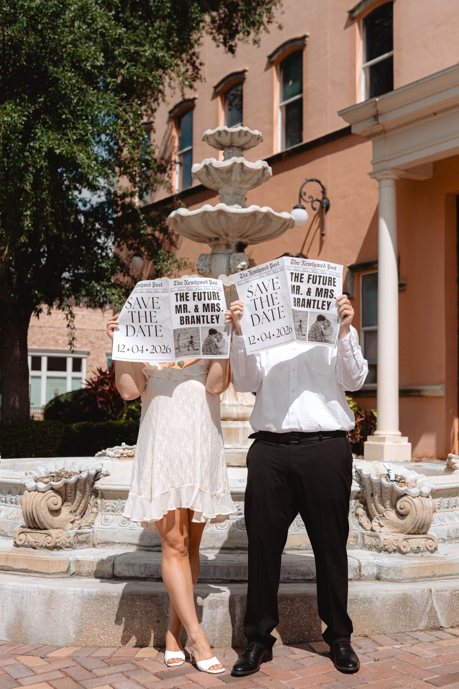 Couple holding Save the Date newspapers in front of a fountain, hiding their faces playfully.
