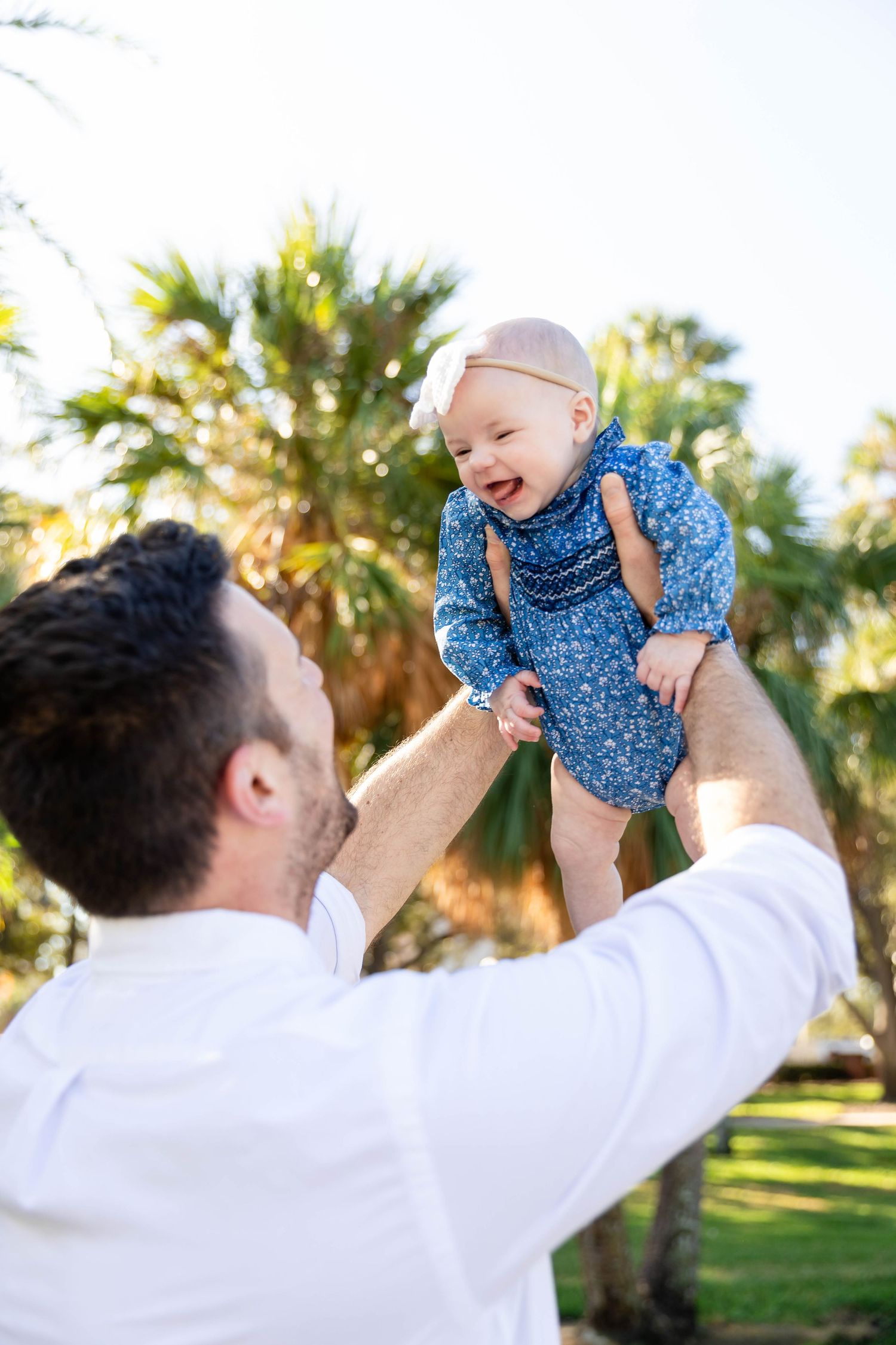 Father joyfully lifts laughing baby girl in blue outfit against sunny palm tree background at the University of Tampa..