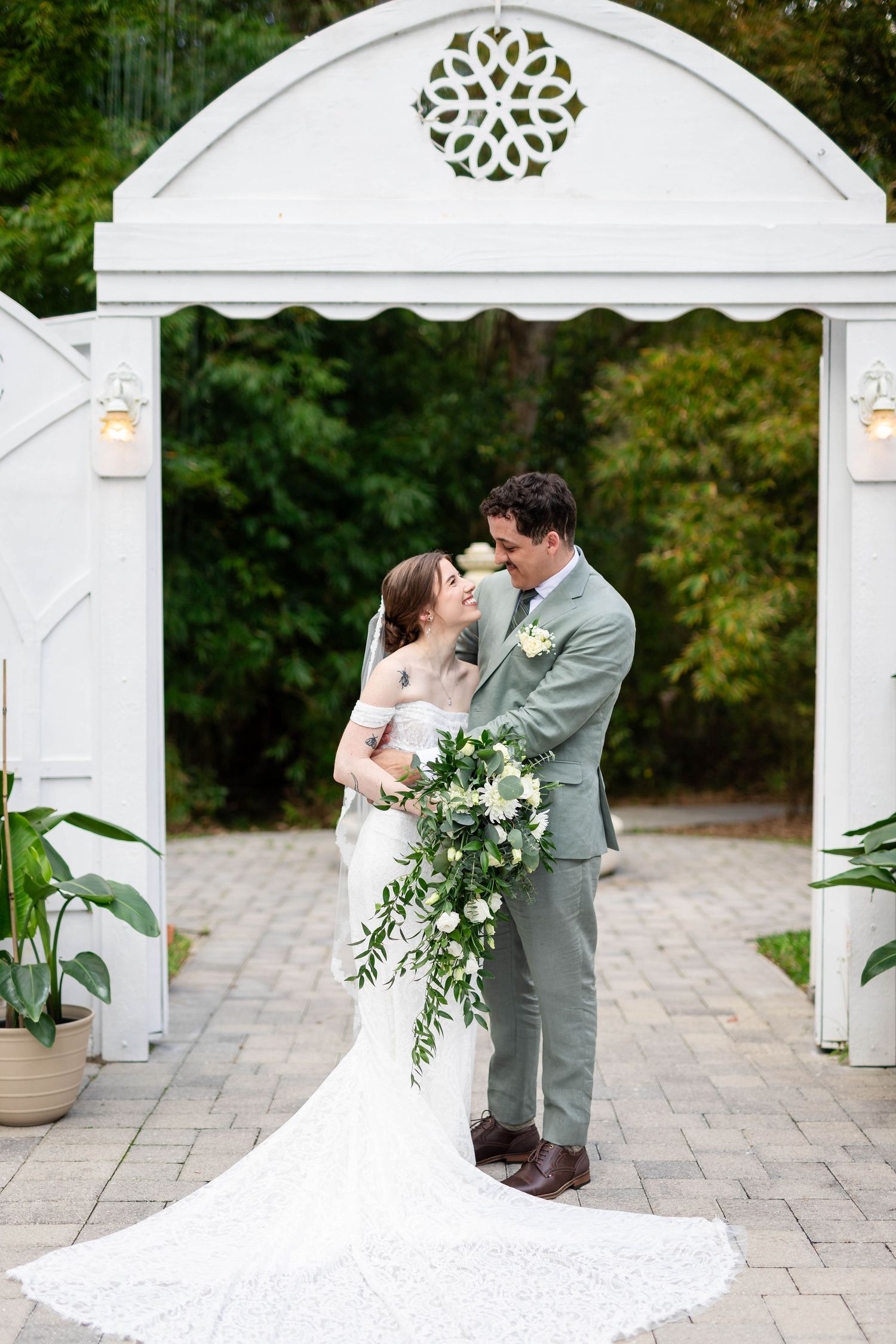 Bride and groom share an intimate moment under a white garden arch surrounded by lush greenery on wedding day.