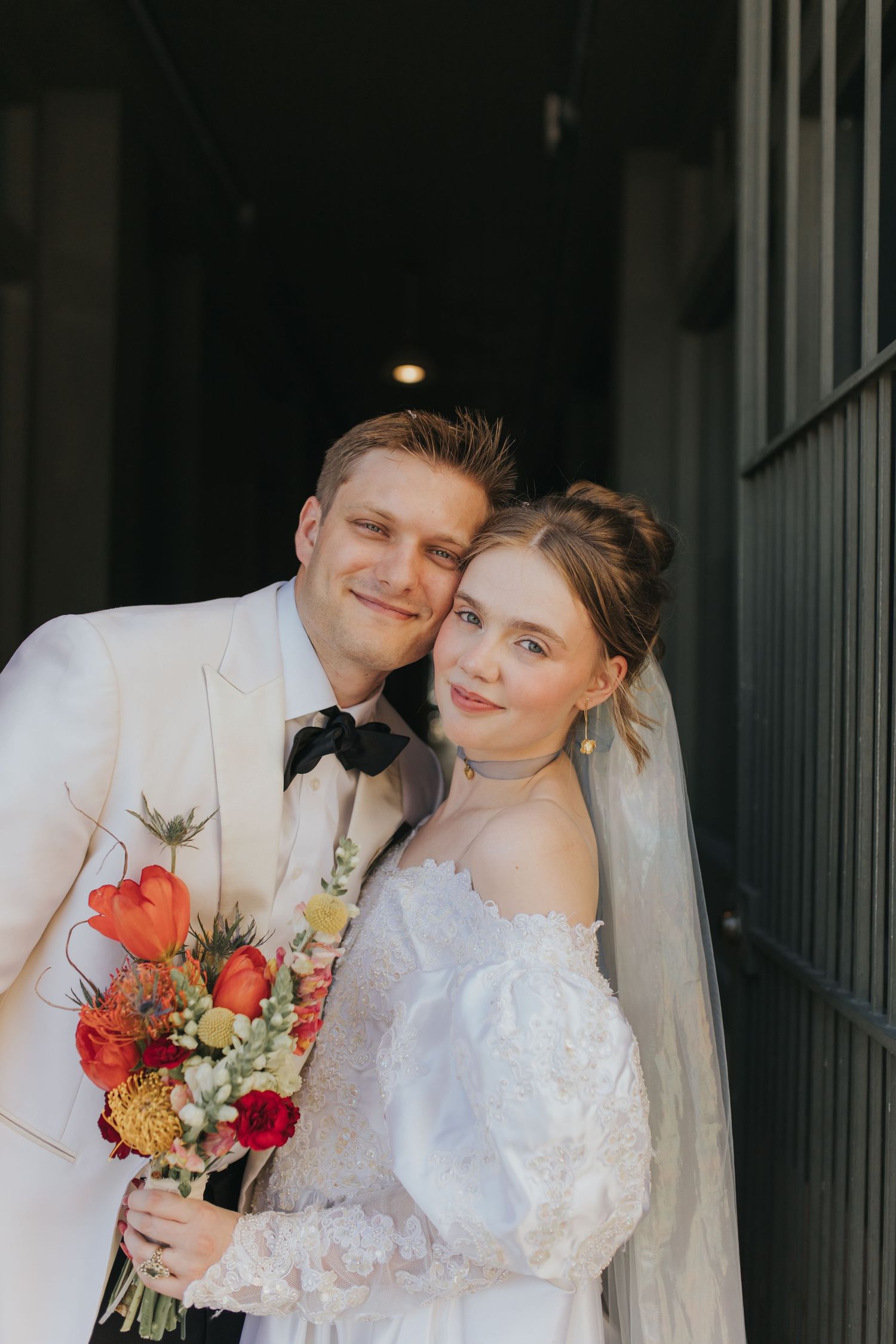 Bride and groom pose with colorful bouquet in white wedding attire outside dark doorway.