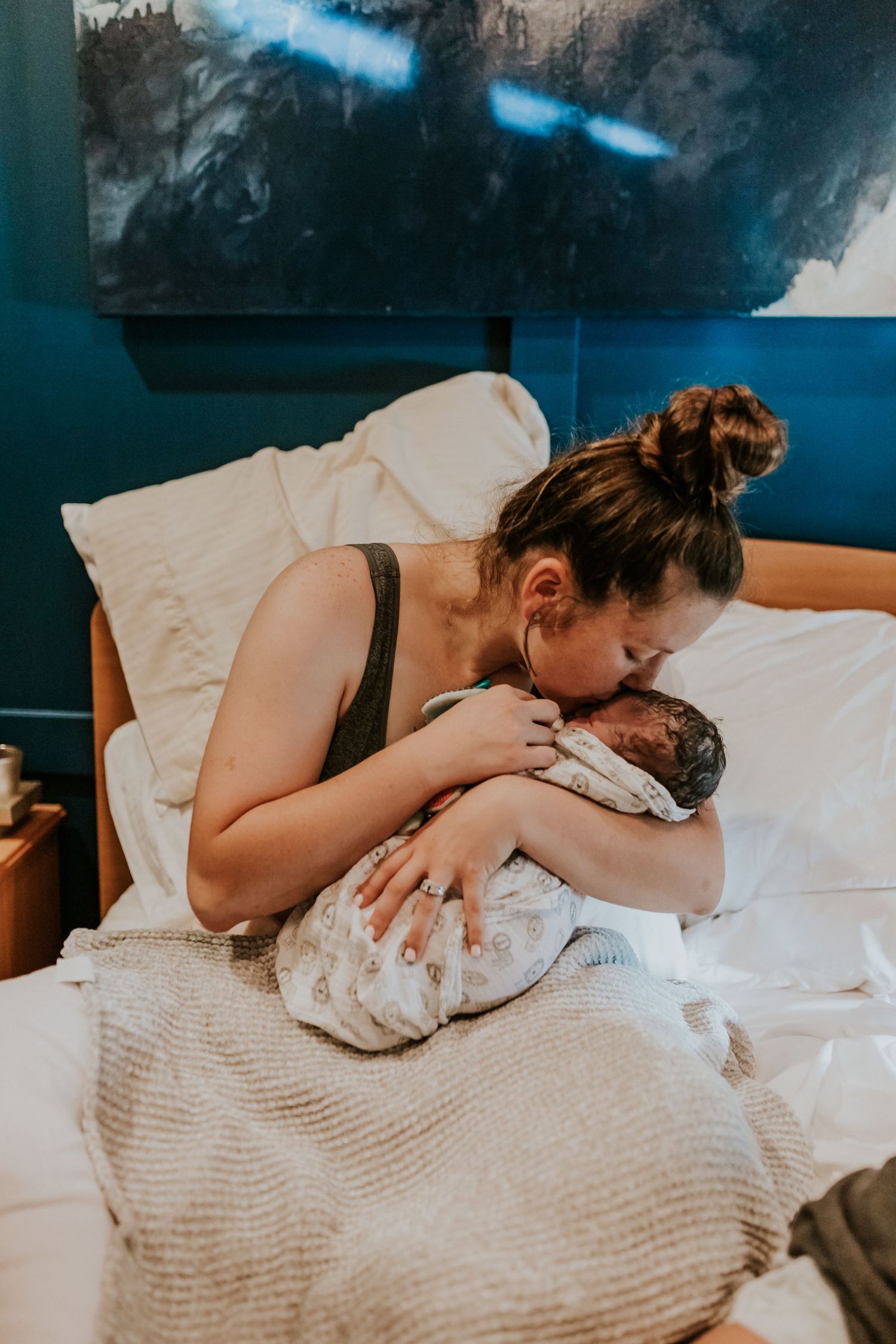 A new mother sits in bed kissing her newborn baby in a tender fresh 48 hospital portrait.