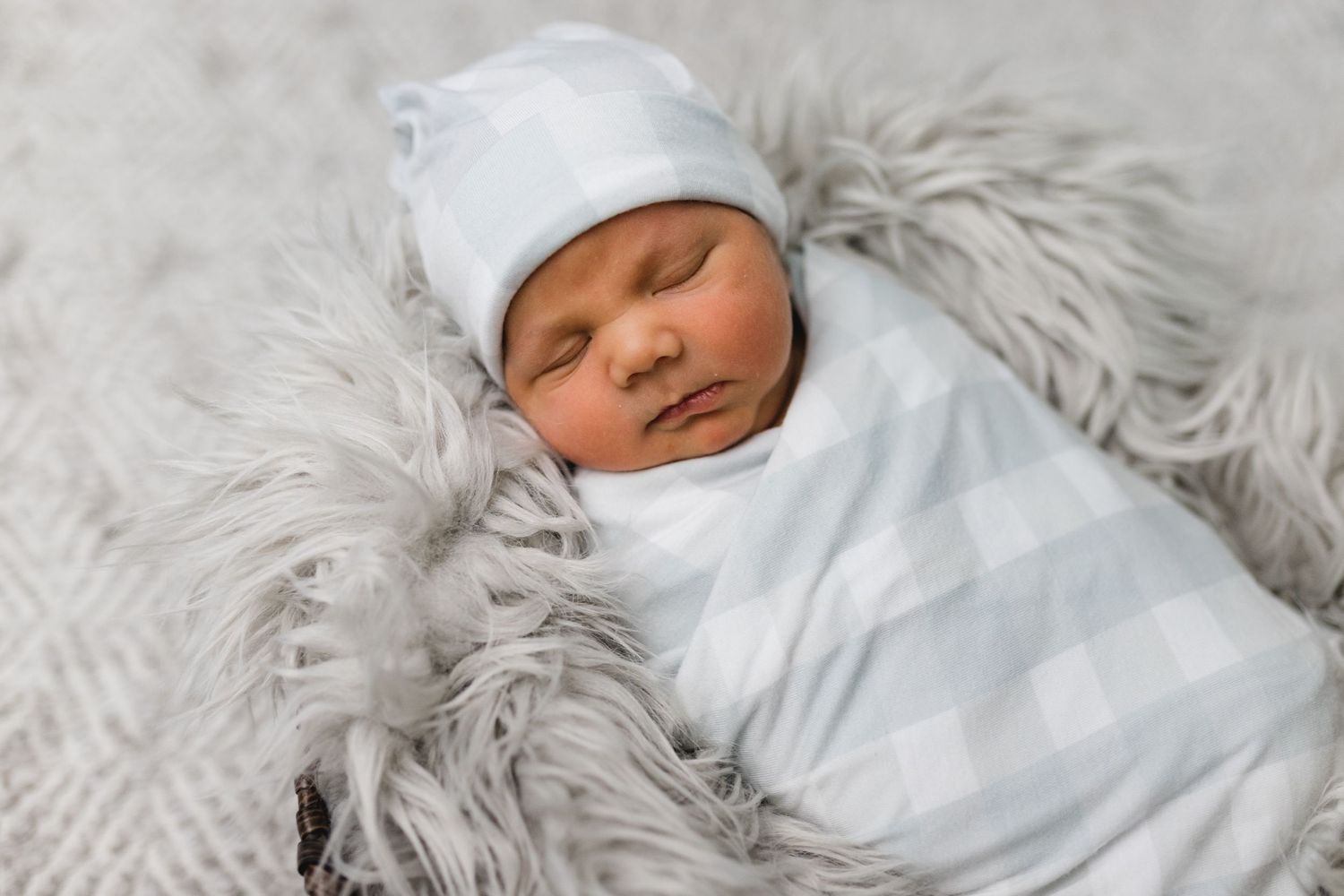 Sleeping newborn baby swaddled in blue plaid wrap resting on gray fluffy blanket wearing white hat.