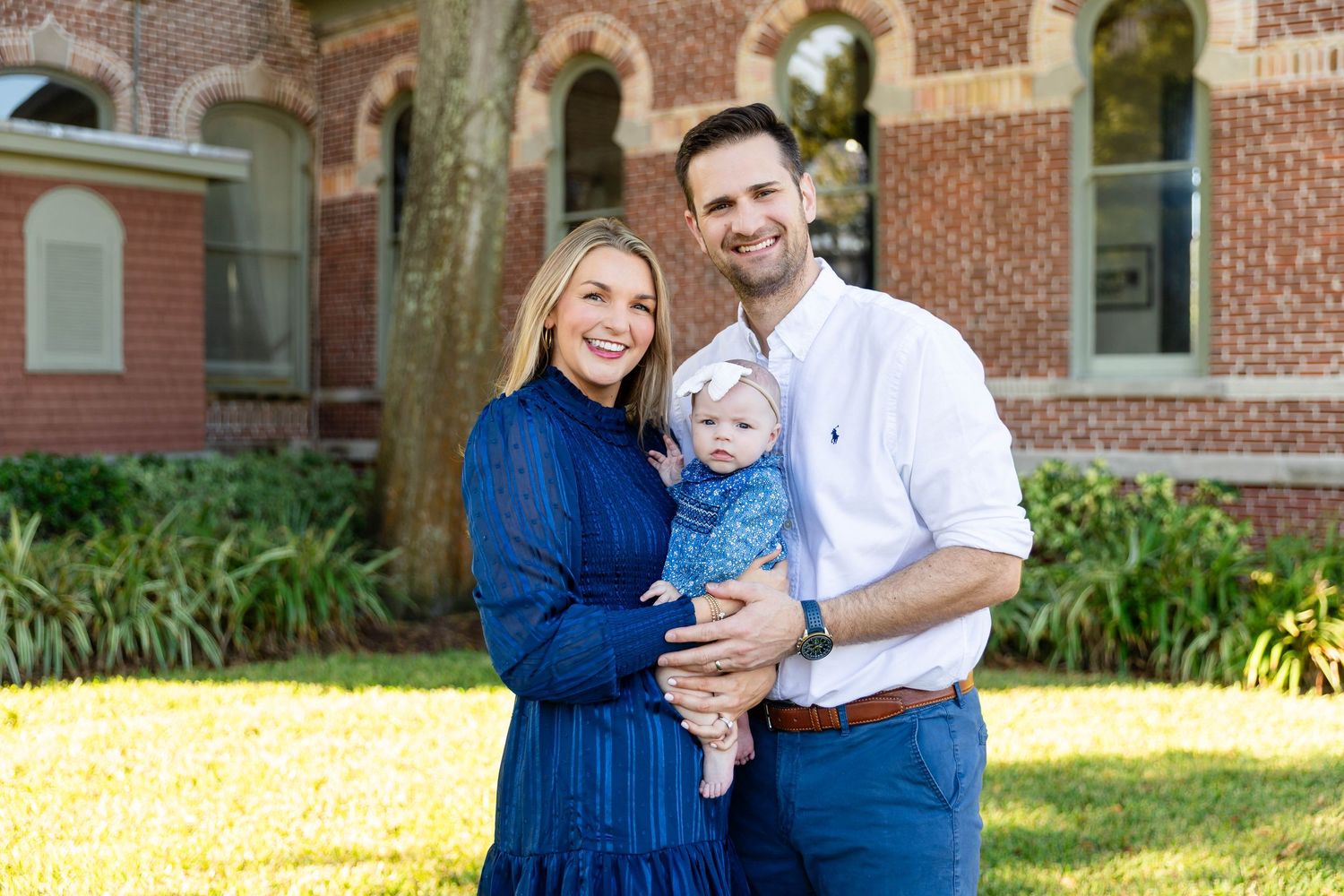 Smiling family of three poses outdoors with baby in blue outfit before historic brick building at the University of Tampa..
