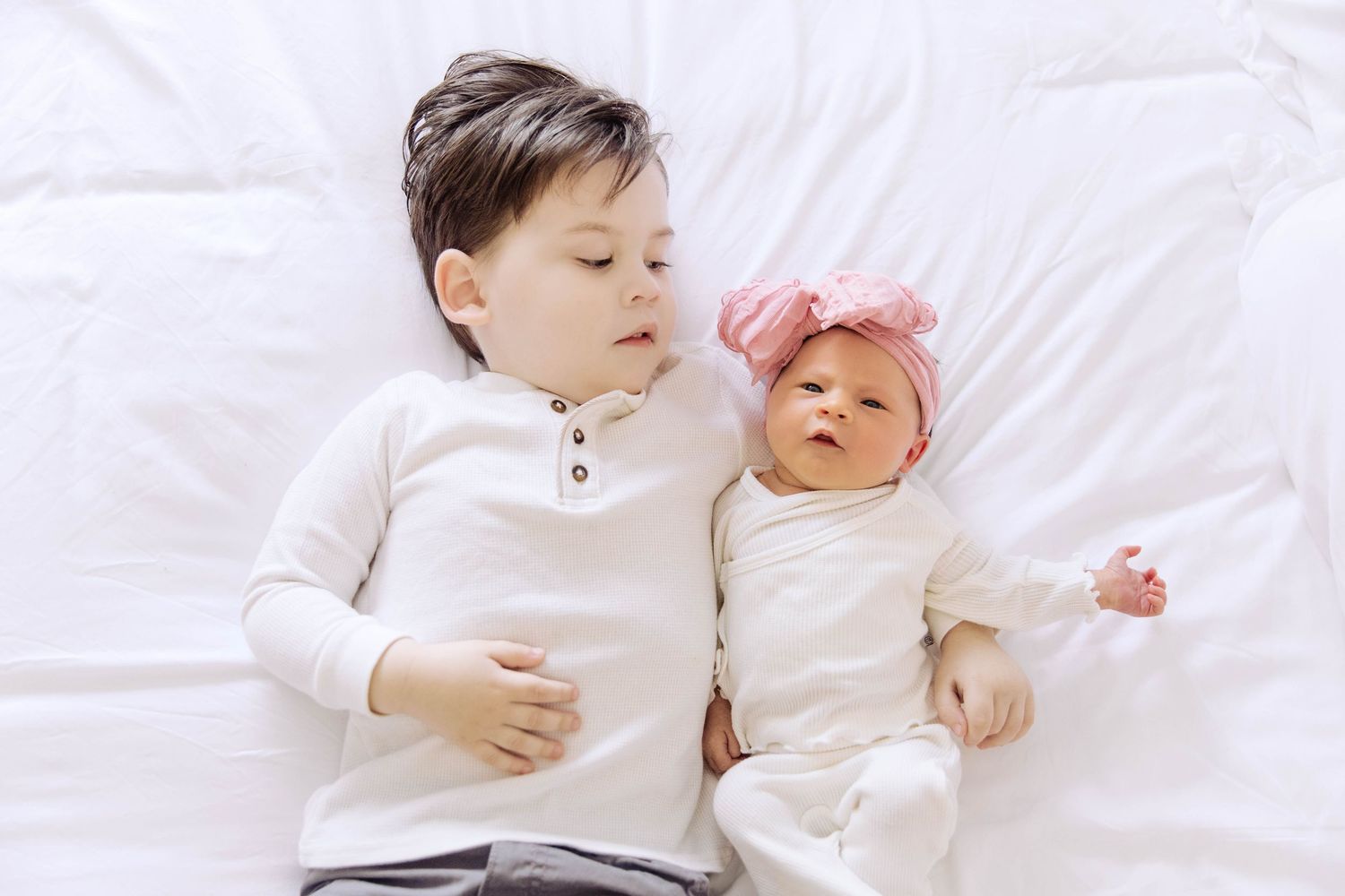 Young boy in white shirt lying next to newborn baby sister with pink bow headband on white bed.