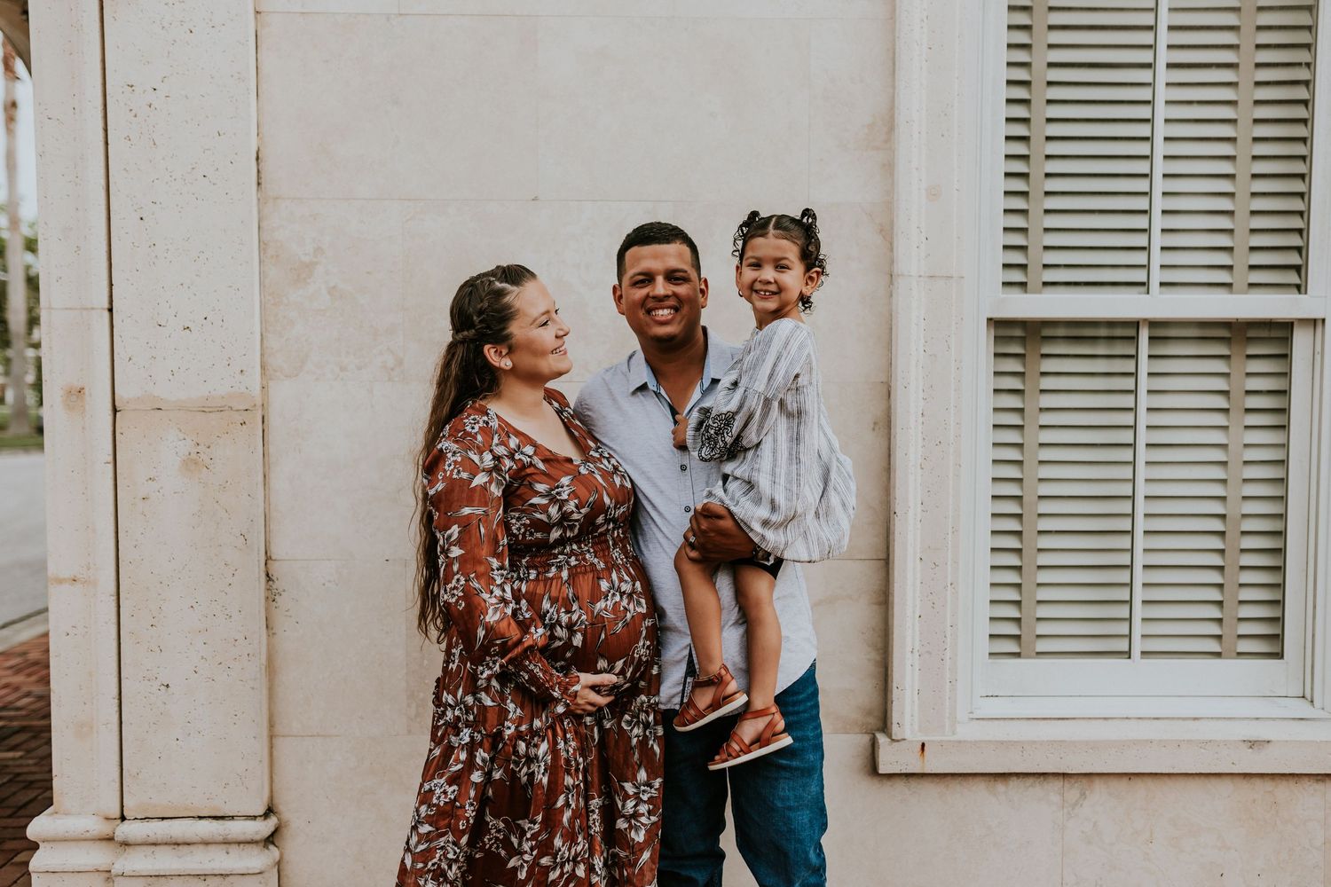 A smiling pregnant woman and man hold their young daughter outside a white building in a family portrait.