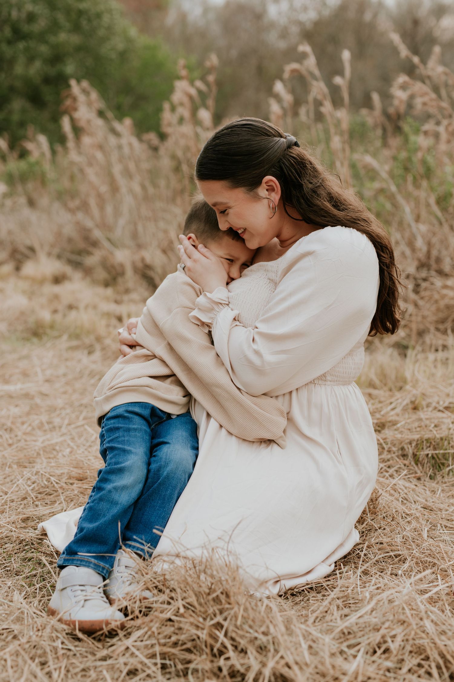 Tender moment as mother in white dress sits in dry grass hugging her young son.