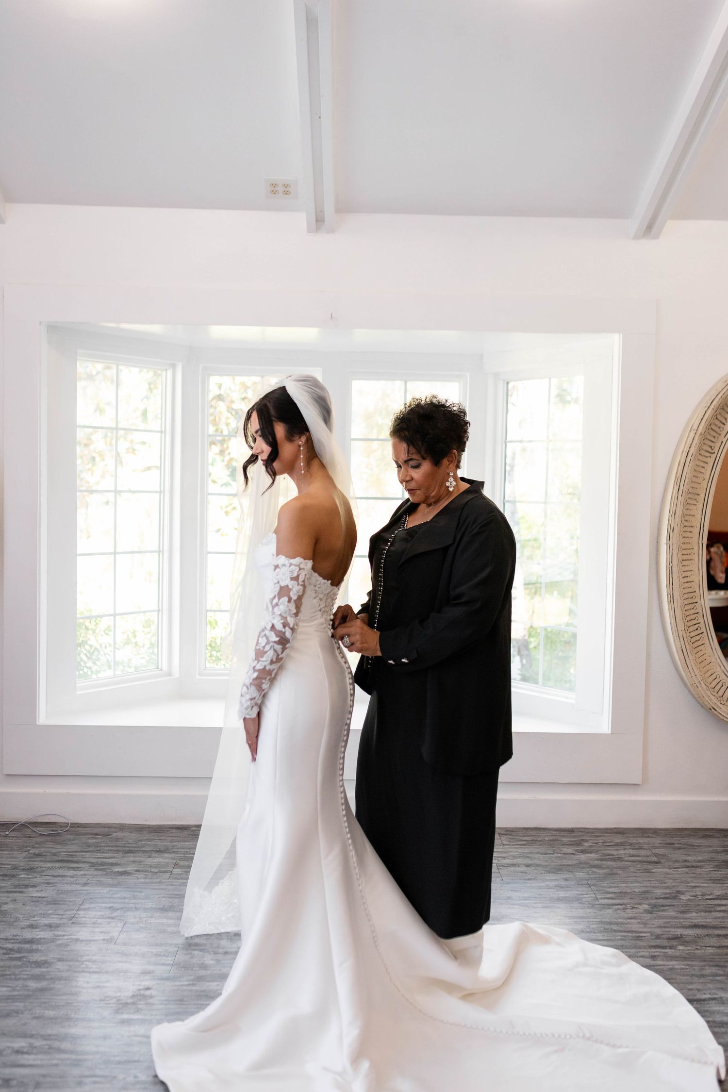 Mother helps bride button her elegant white lace wedding gown in a bright, sunlit bridal suite in Tampa.