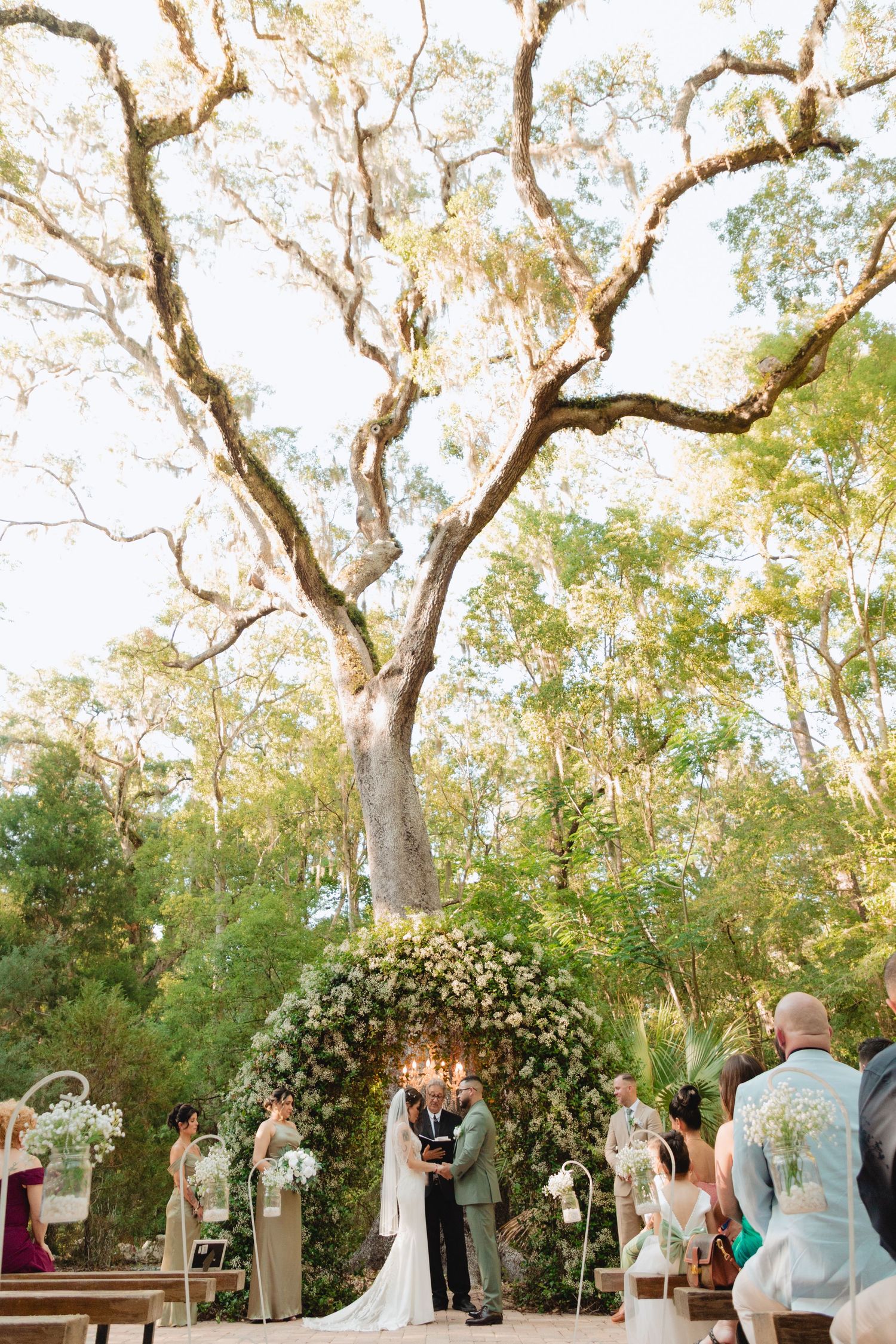 Bride and groom exchange vows under a white floral arch beneath a grand oak tree in outdoor ceremony.