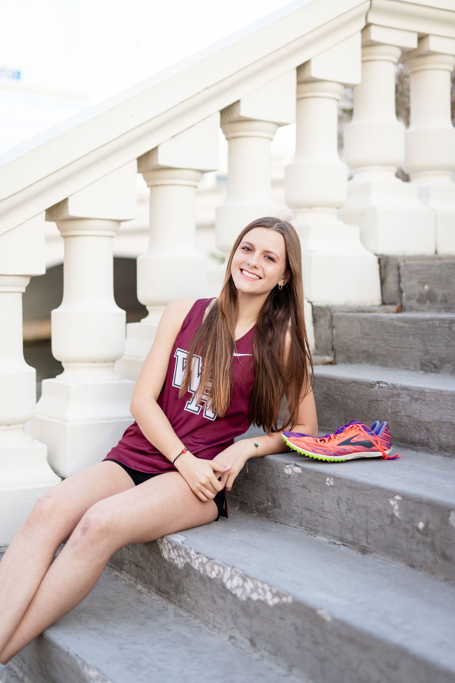 Smiling teen girl in maroon track jersey sitting on stone steps with colorful running shoes at the University of Tampa.