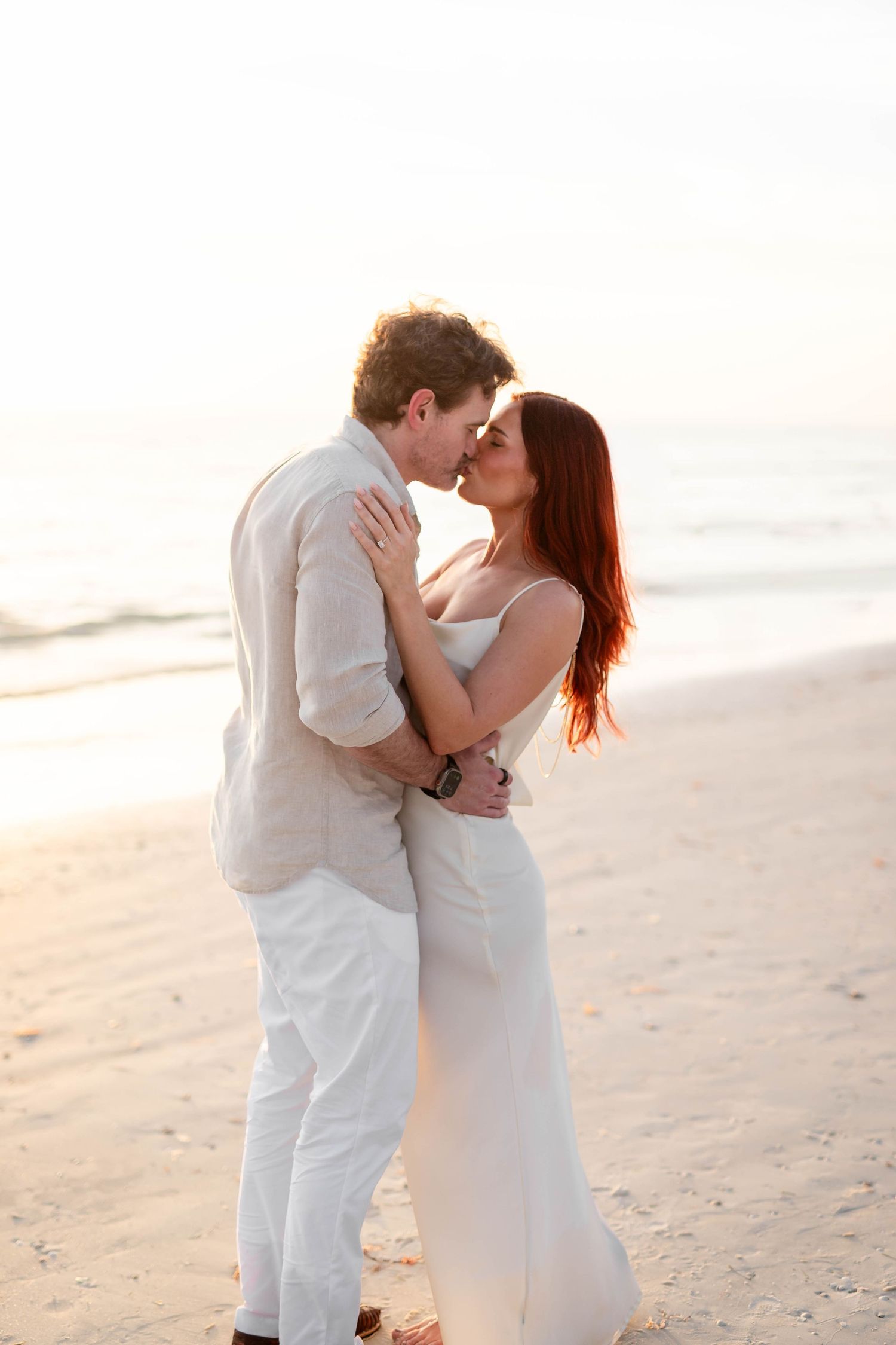 Couple embracing and kissing on a sunlit beach at sunset, woman in white dress, man in linen at Honeymoon Island.