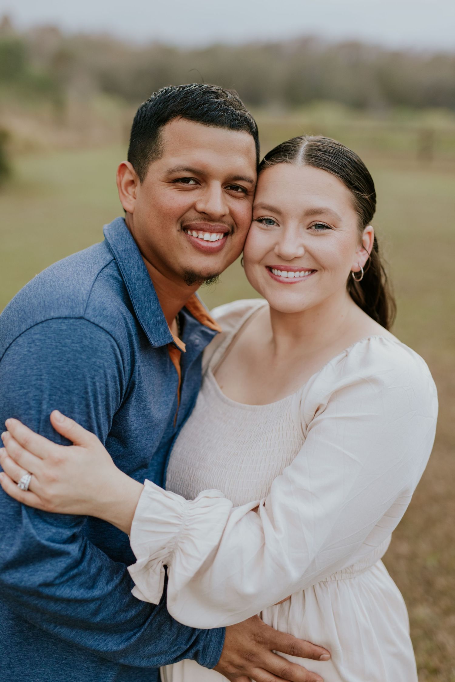 Smiling couple embracing outdoors, woman in cream dress and man in blue shirt.