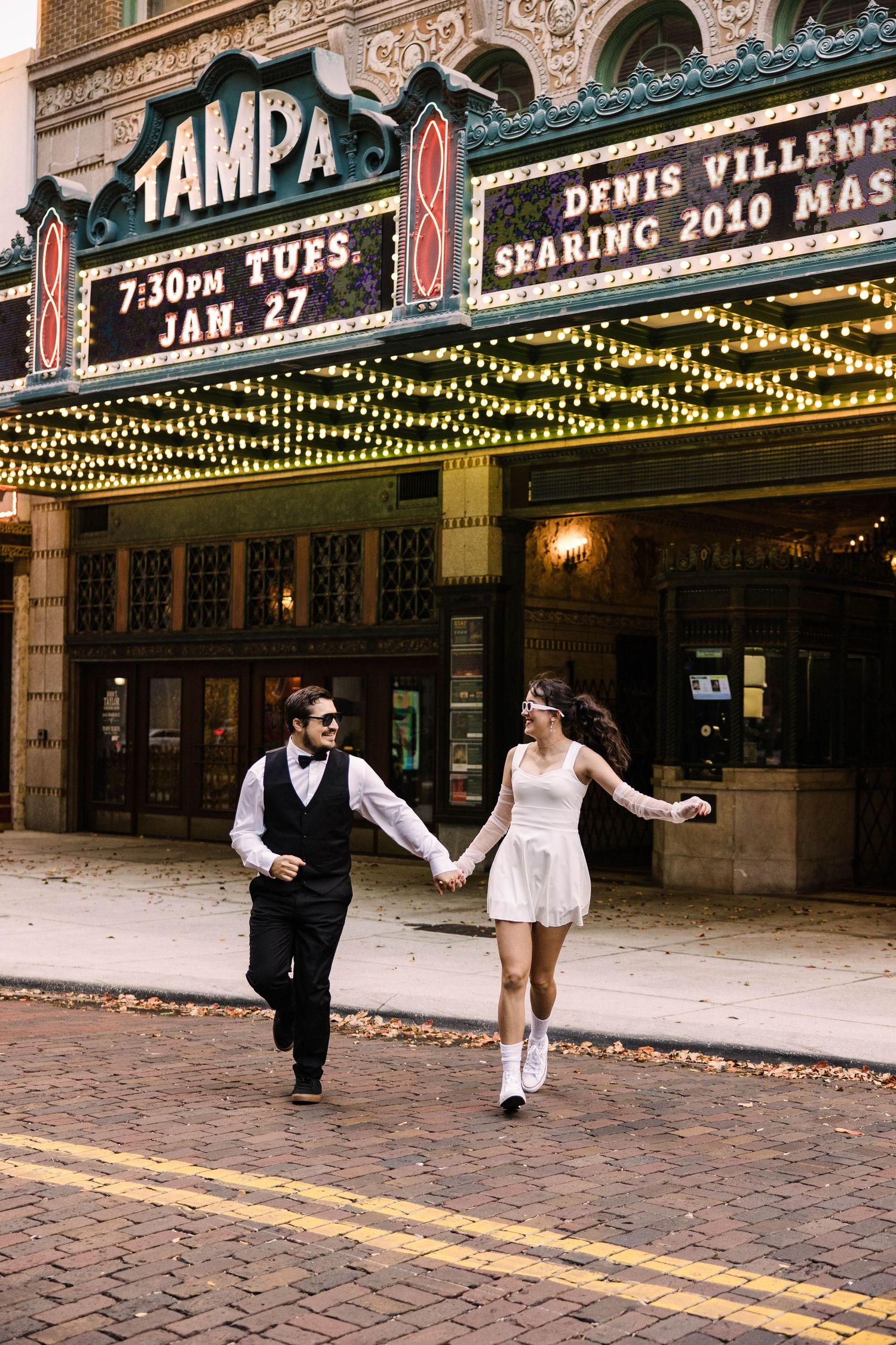 Bride and groom run hand in hand past the iconic Tampa Theatre marquee at night.