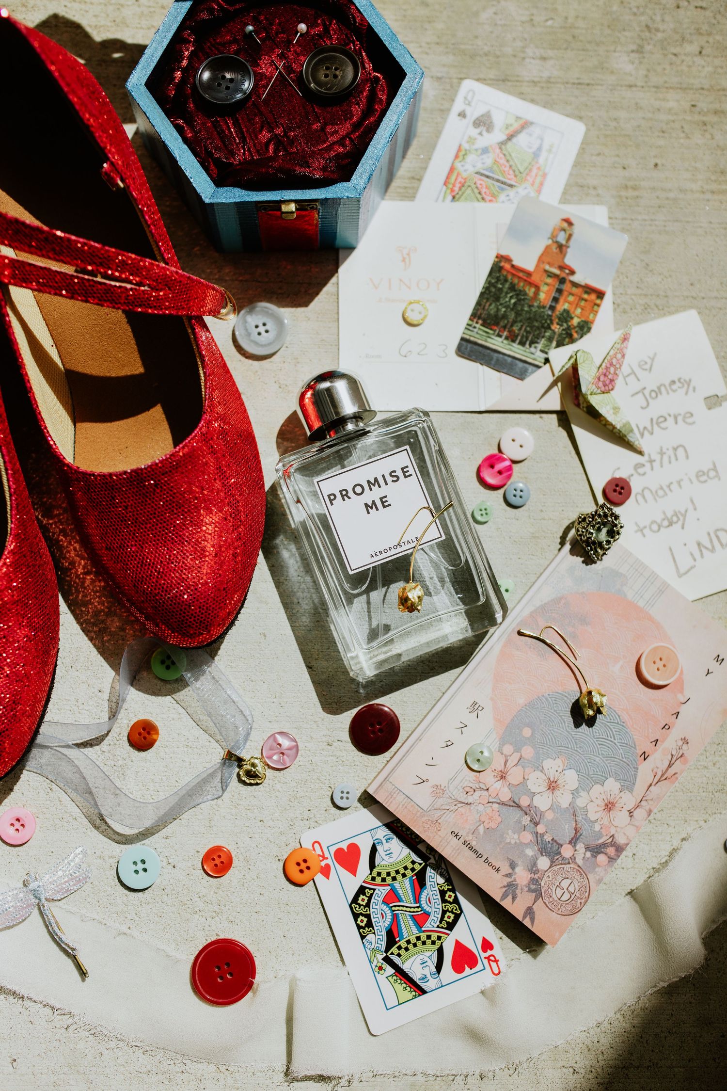 Red glitter heels, Promise Me perfume, playing cards, colorful buttons, and jewelry arranged as wedding flat lay accessories.