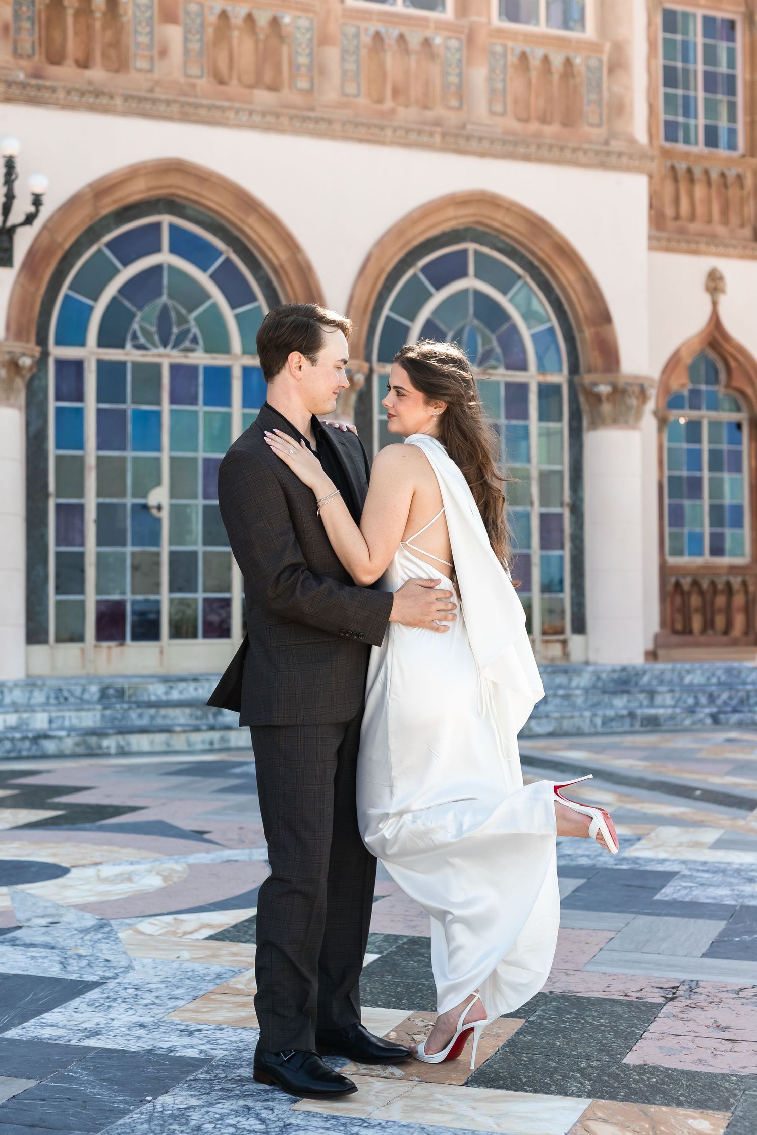 Bride in backless white gown and groom embrace before ornate stained glass windows at wedding venue at the Ringling Museum.