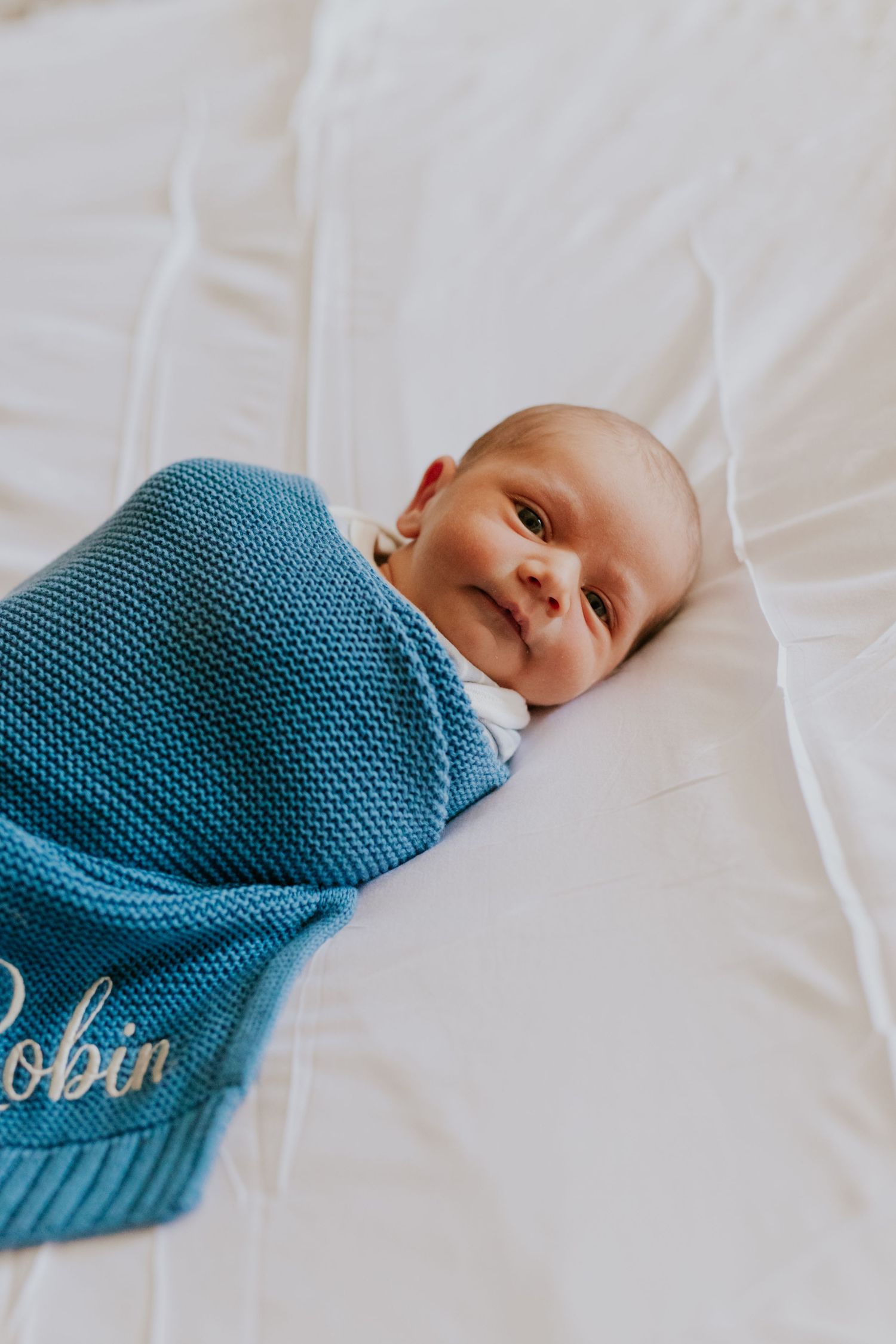 Smiling newborn baby swaddled in a blue knit blanket embroidered with the name Robin.