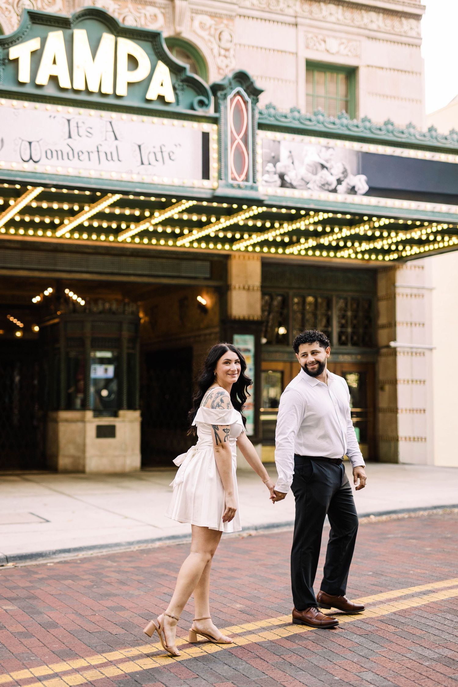 Smiling couple holding hands walking in front of the iconic Tampa Theatre marquee at dusk in Tampa.