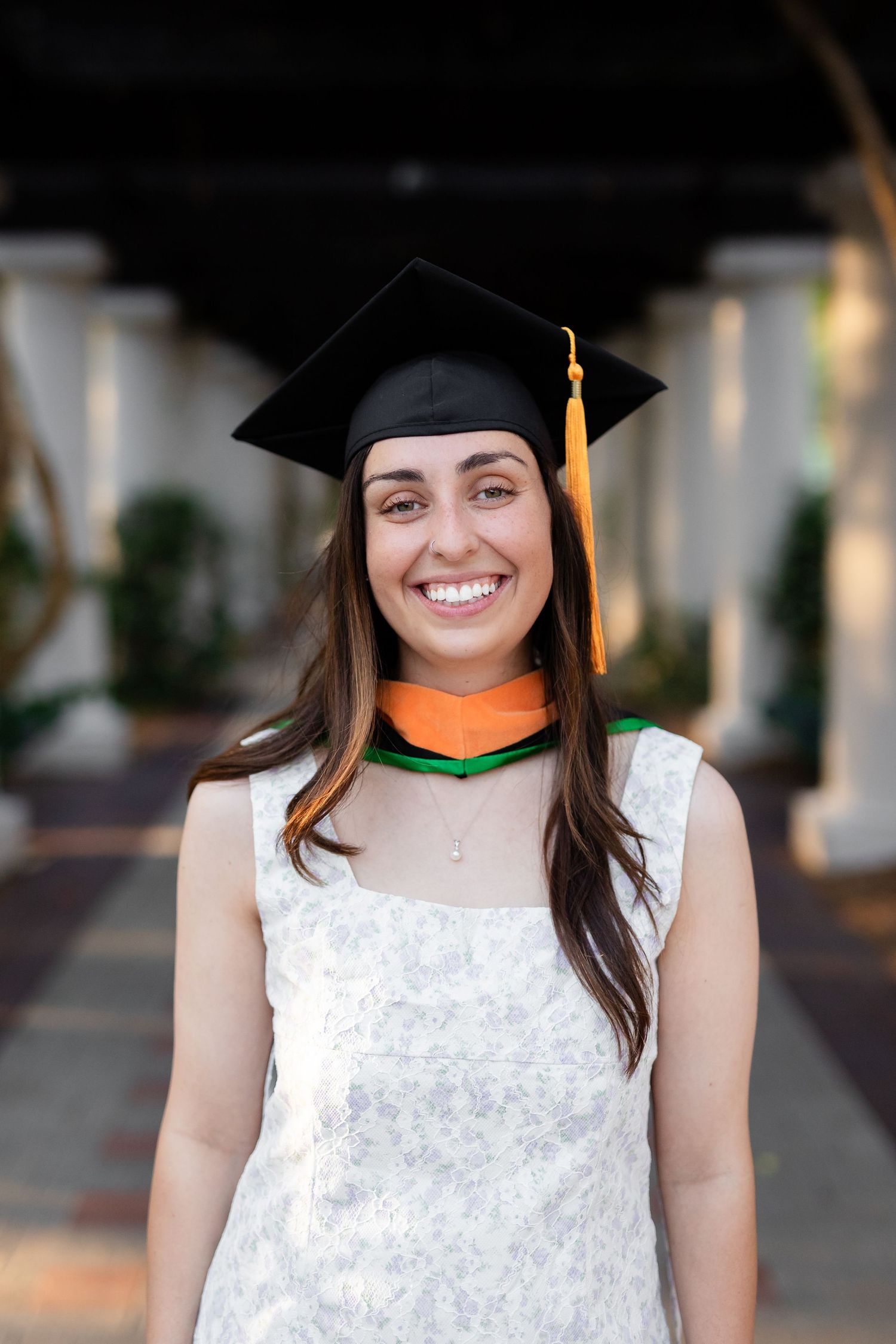 Smiling female graduate wearing black mortarboard cap with gold tassel and white floral dress outdoors at the University of South Florida.