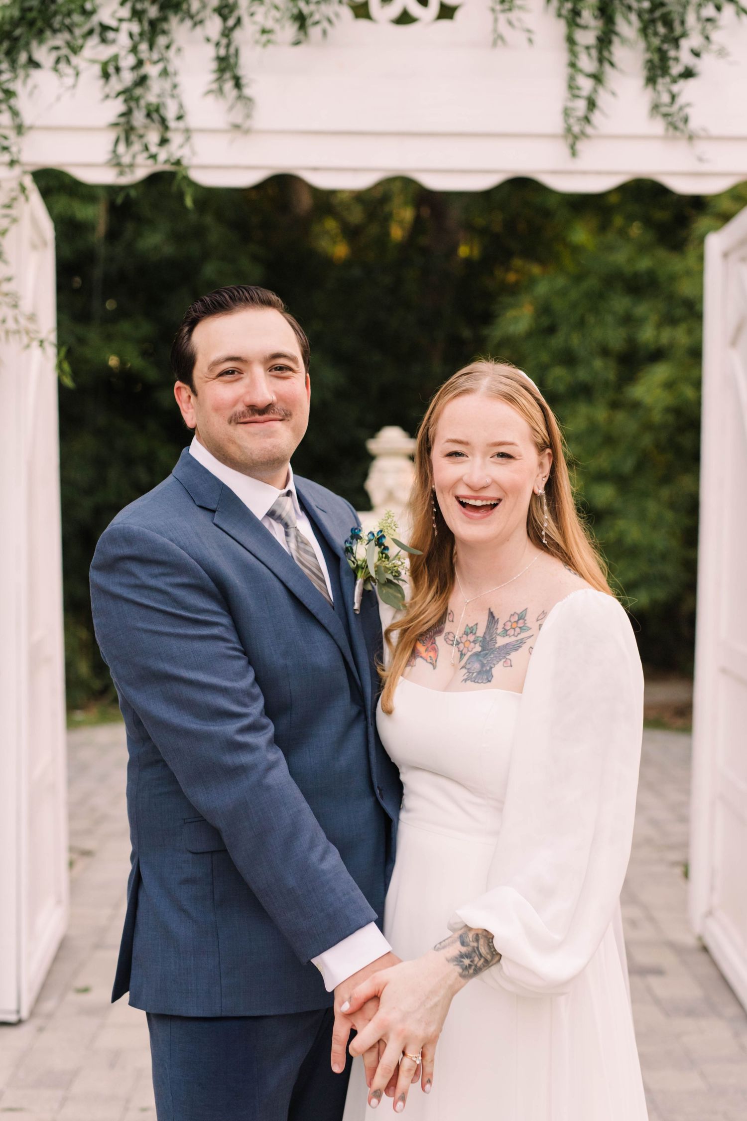 Smiling bride and groom pose together under a white arbor with lush greenery backdrop.