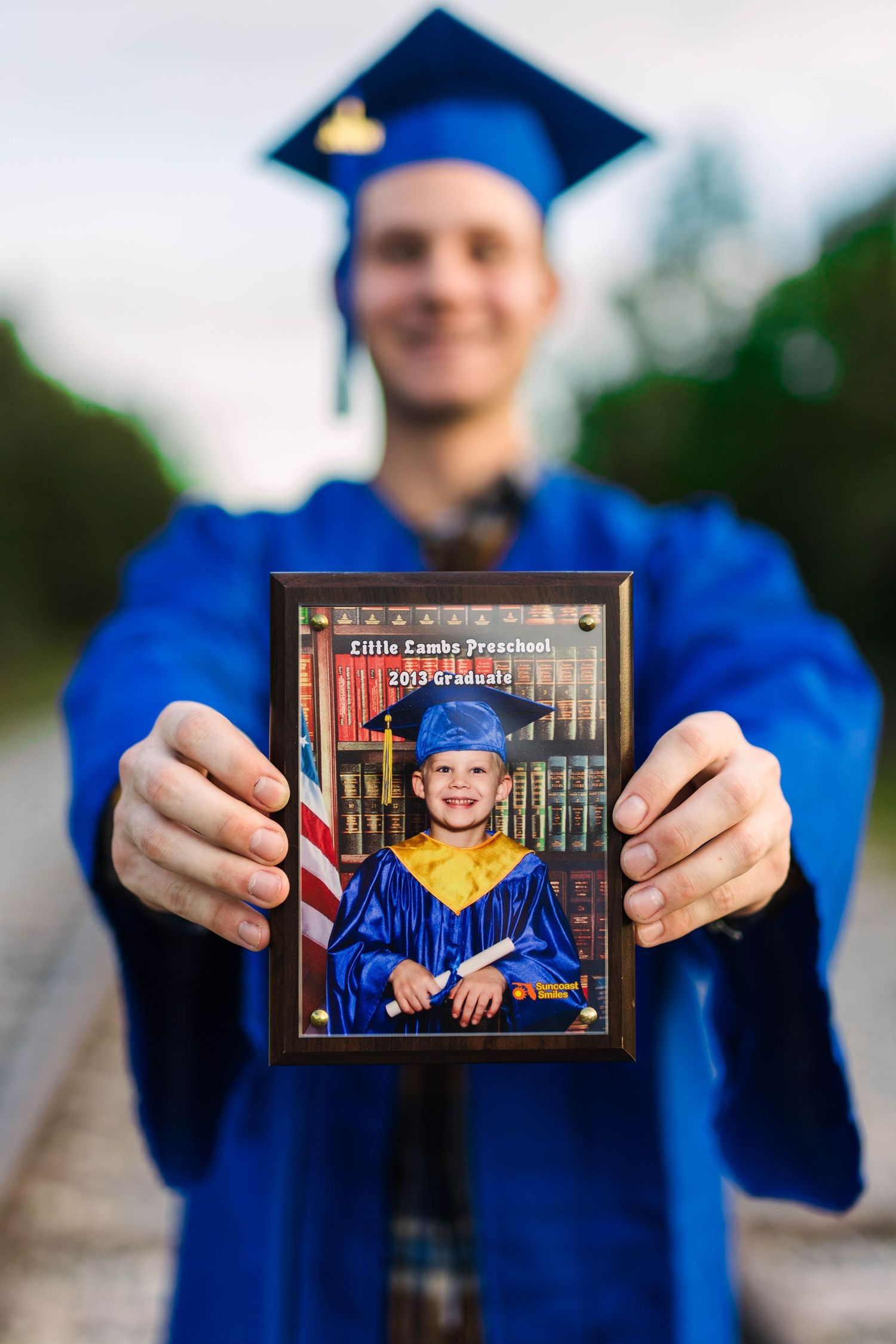 Male graduate in blue cap and gown holding framed preschool graduation photo outdoors.
