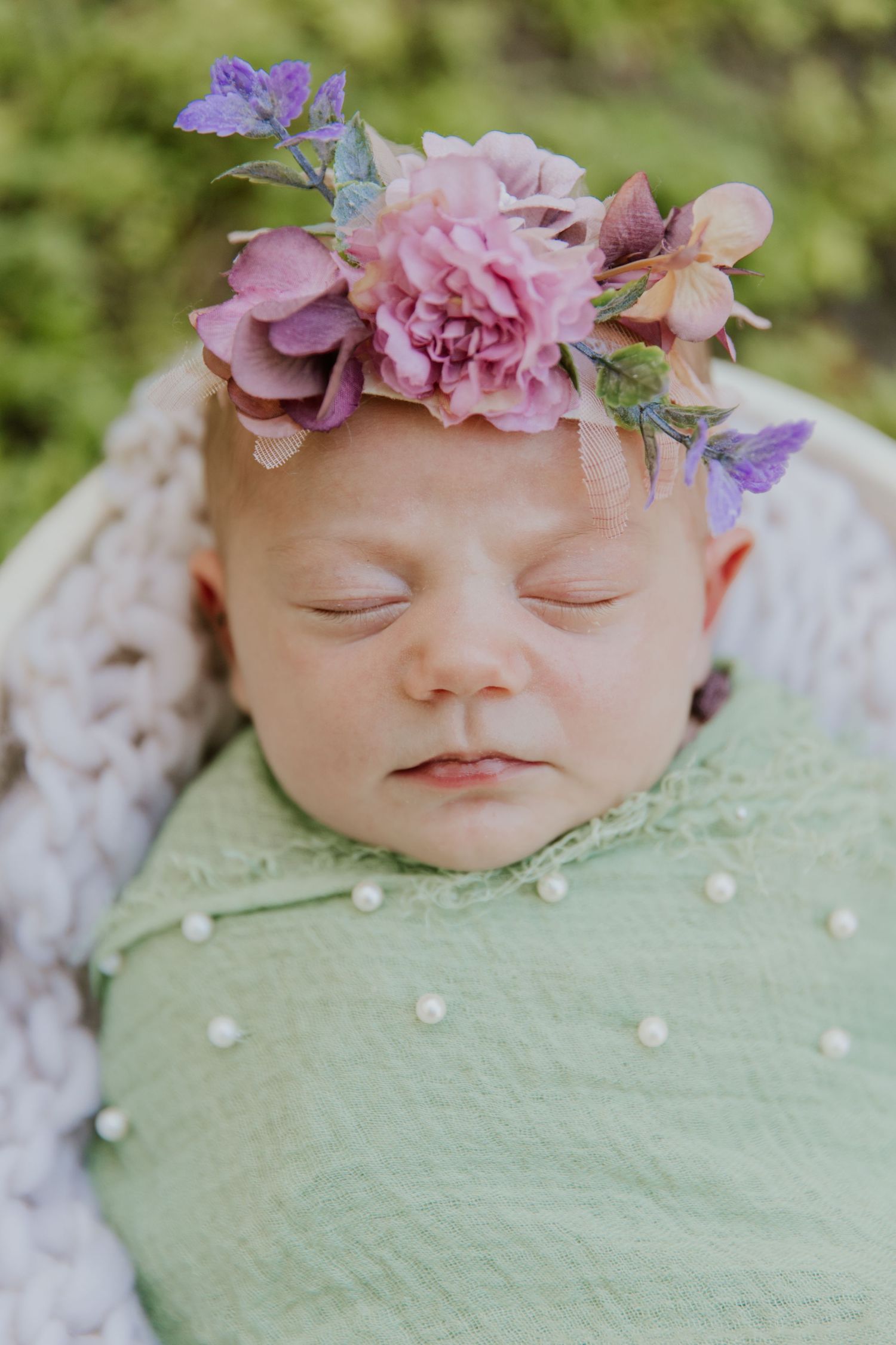 Sleeping newborn baby swaddled in green wrap adorned with a beautiful purple and pink floral crown.