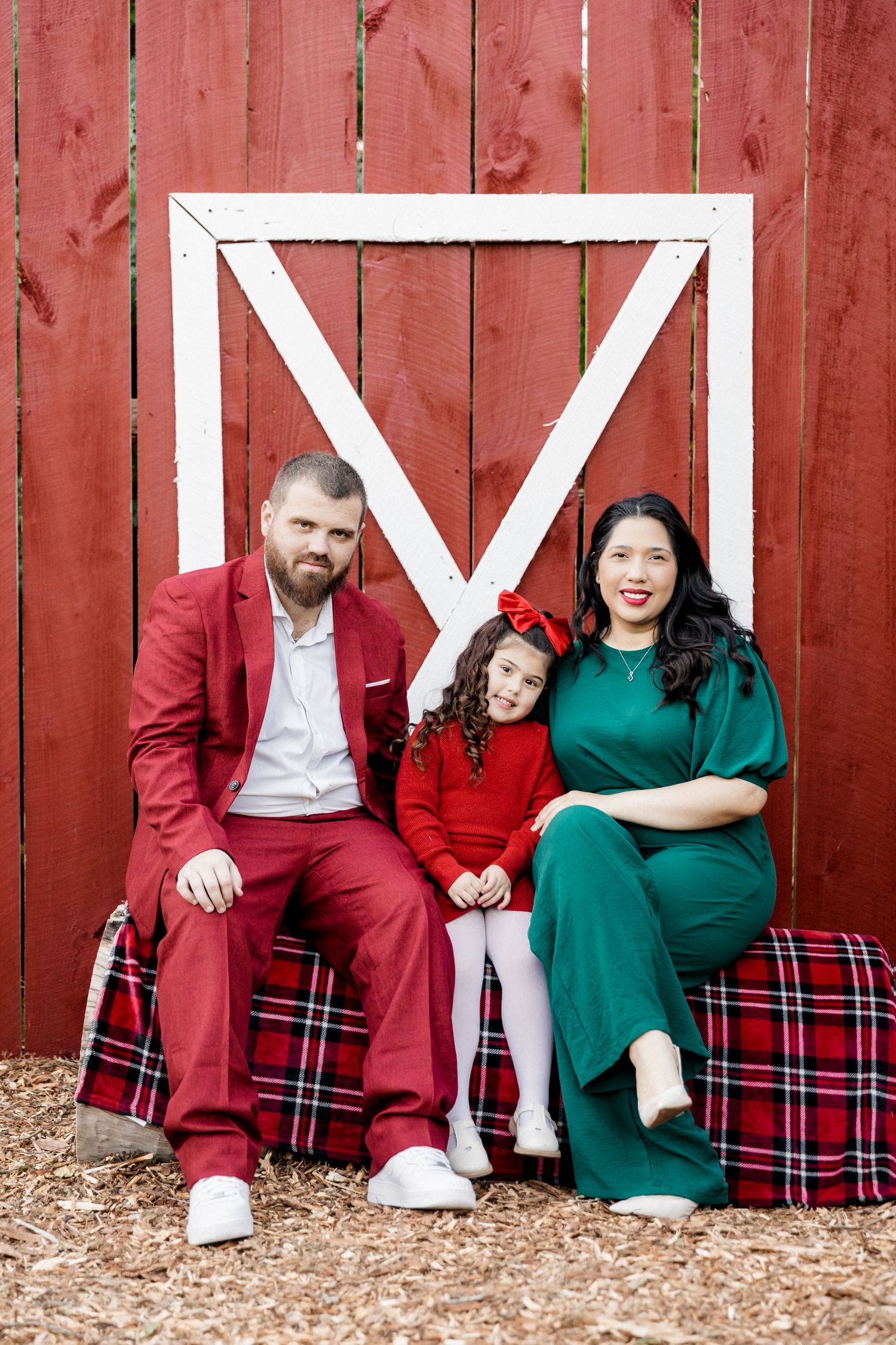 Family of three sits before red barn door, dressed in festive red and green holiday attire.