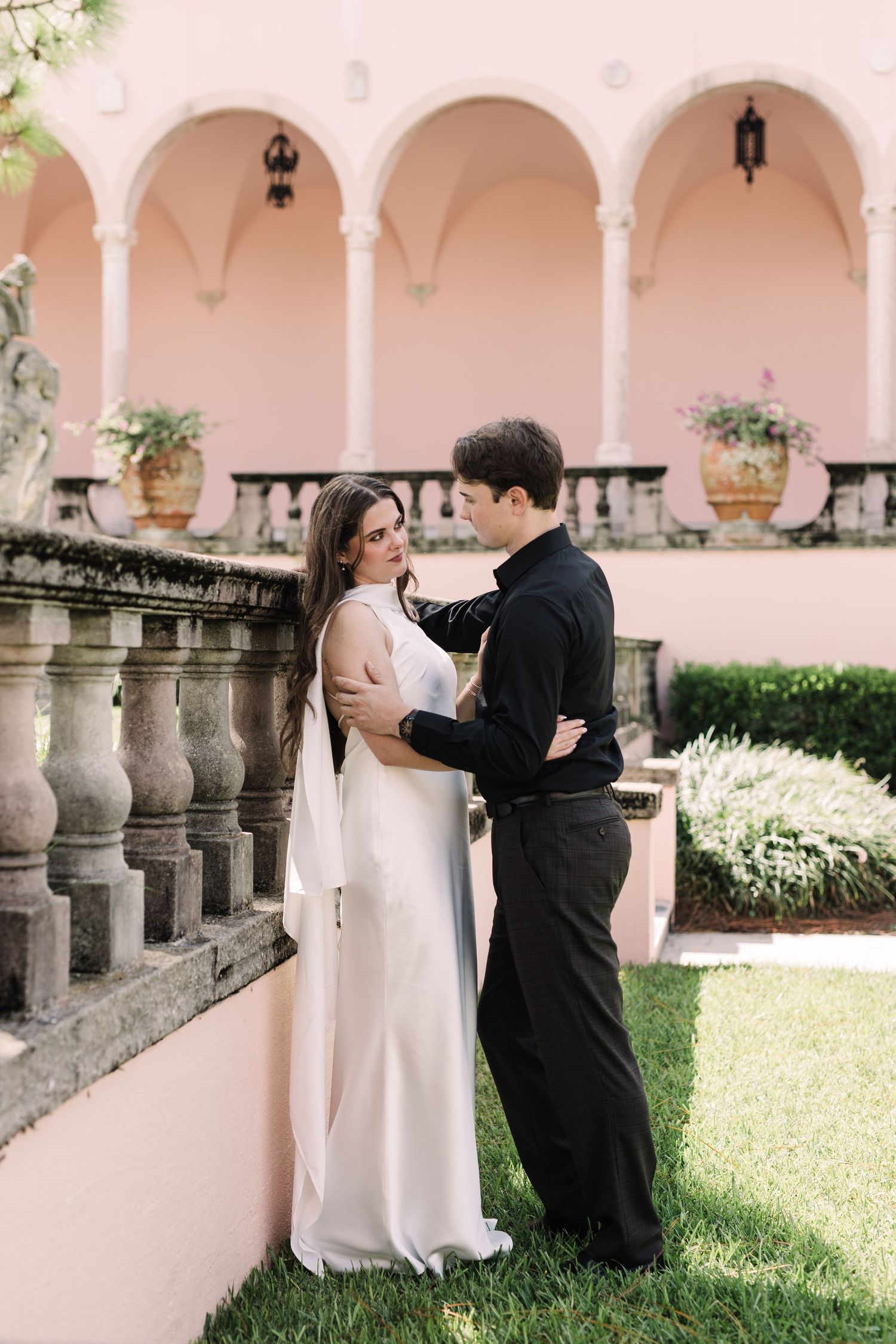 Bride in white gown with veil and groom in black suit embrace in elegant pink courtyard garden at the Ringling Museum.