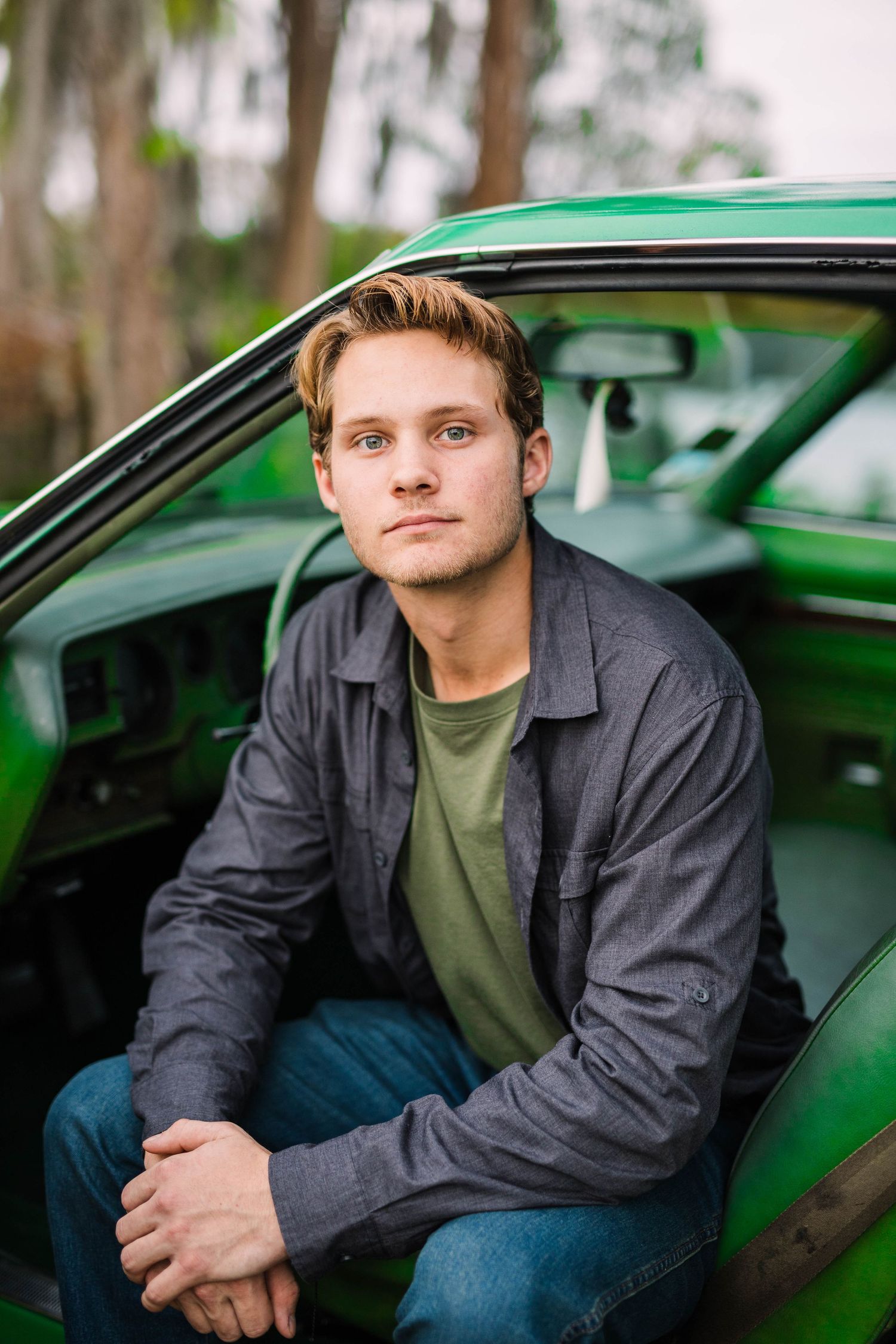 Handsome young man with blue eyes leaning against vintage green car outdoors at dusk.
