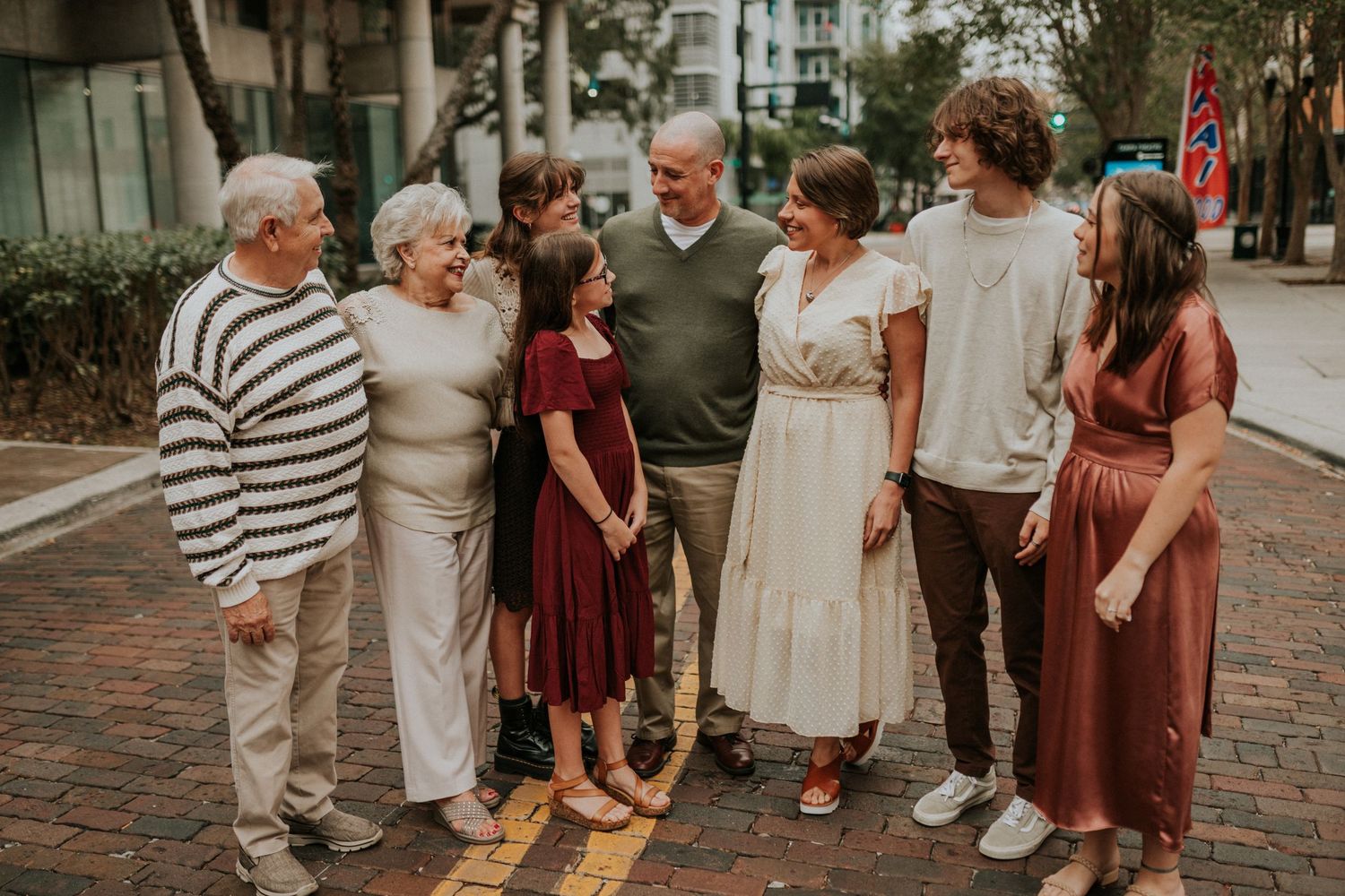 Multigenerational family of seven smiles together on a brick sidewalk in an urban setting.