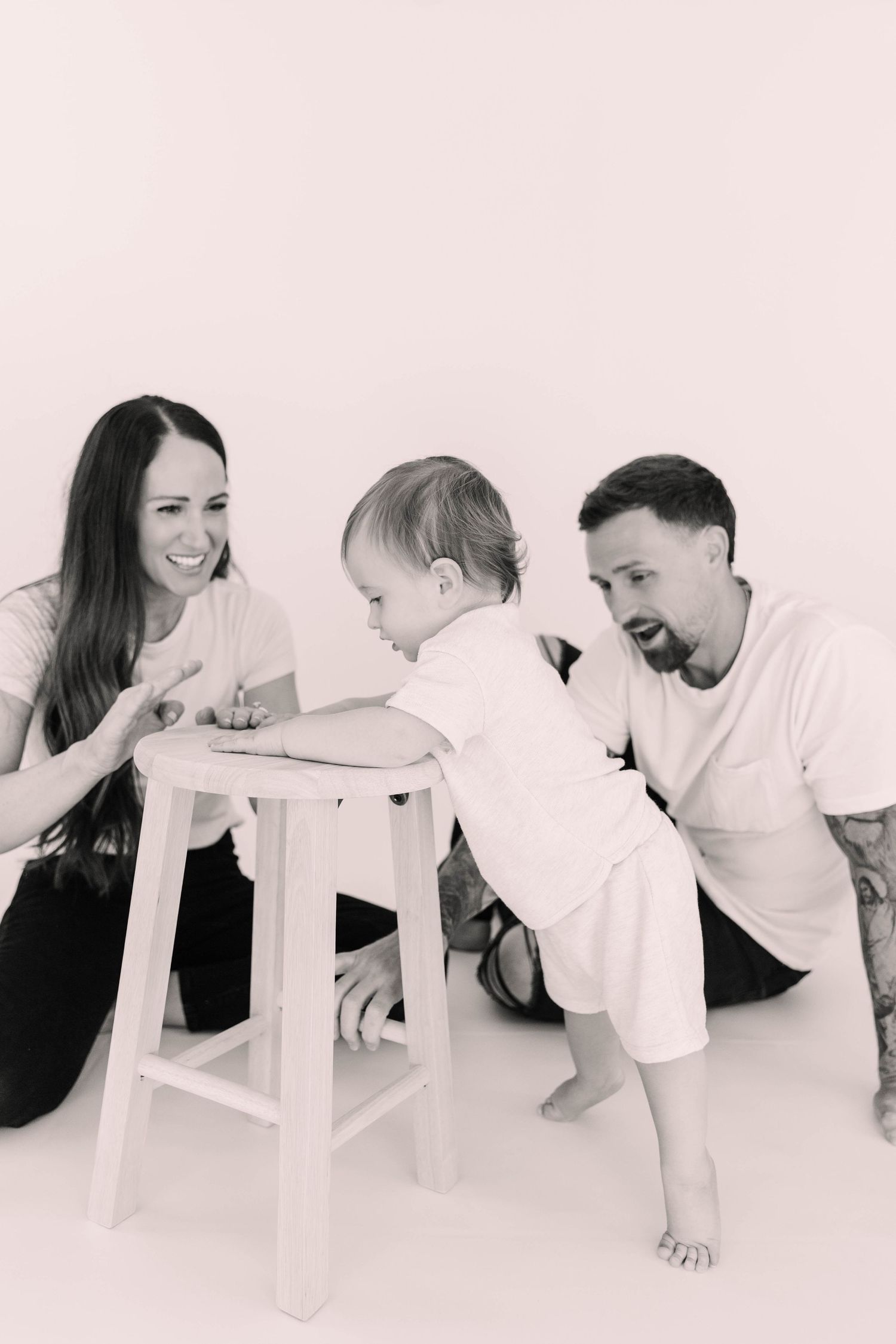 Black and white photo of parents smiling at their baby standing at a wooden stool.