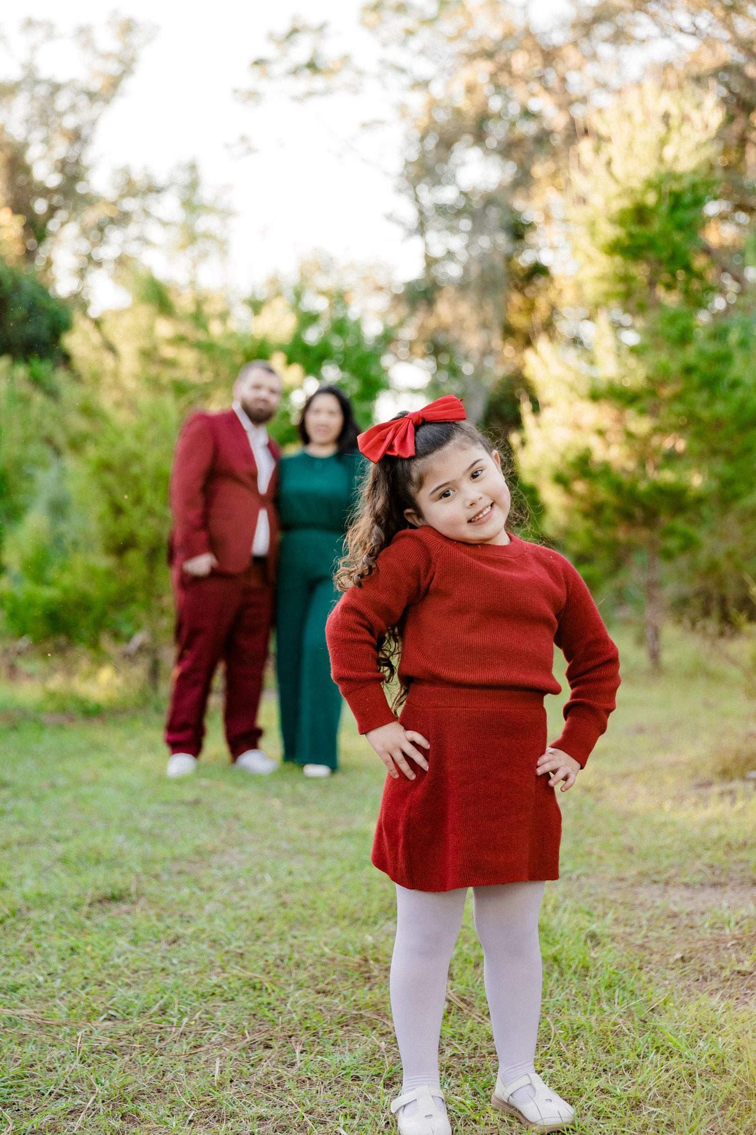 Young girl in red dress poses confidently with parents blurred in background at tree farm.