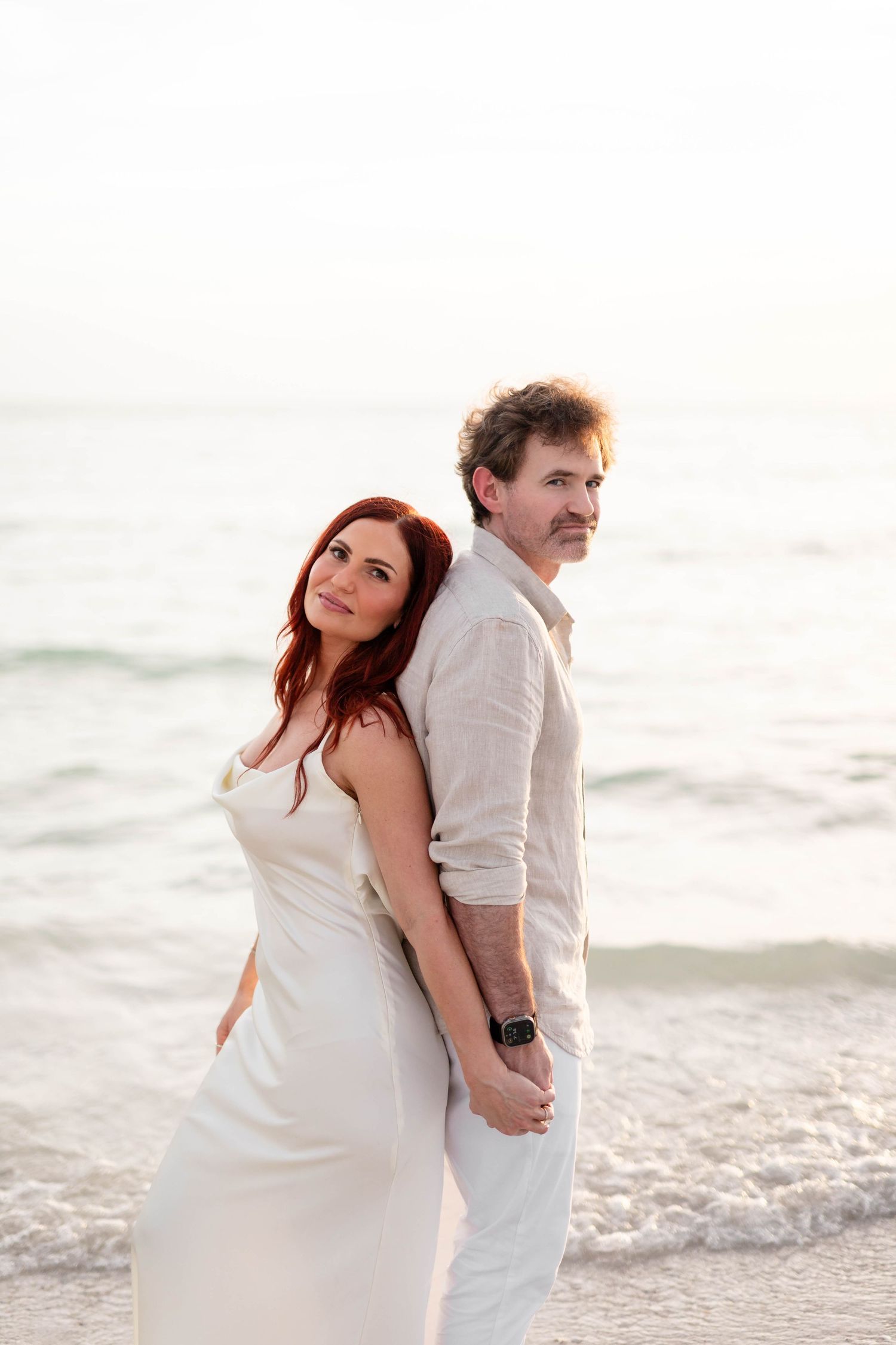 Couple in white attire stands back-to-back on beach at sunset by ocean waves at Honeymoon Island.