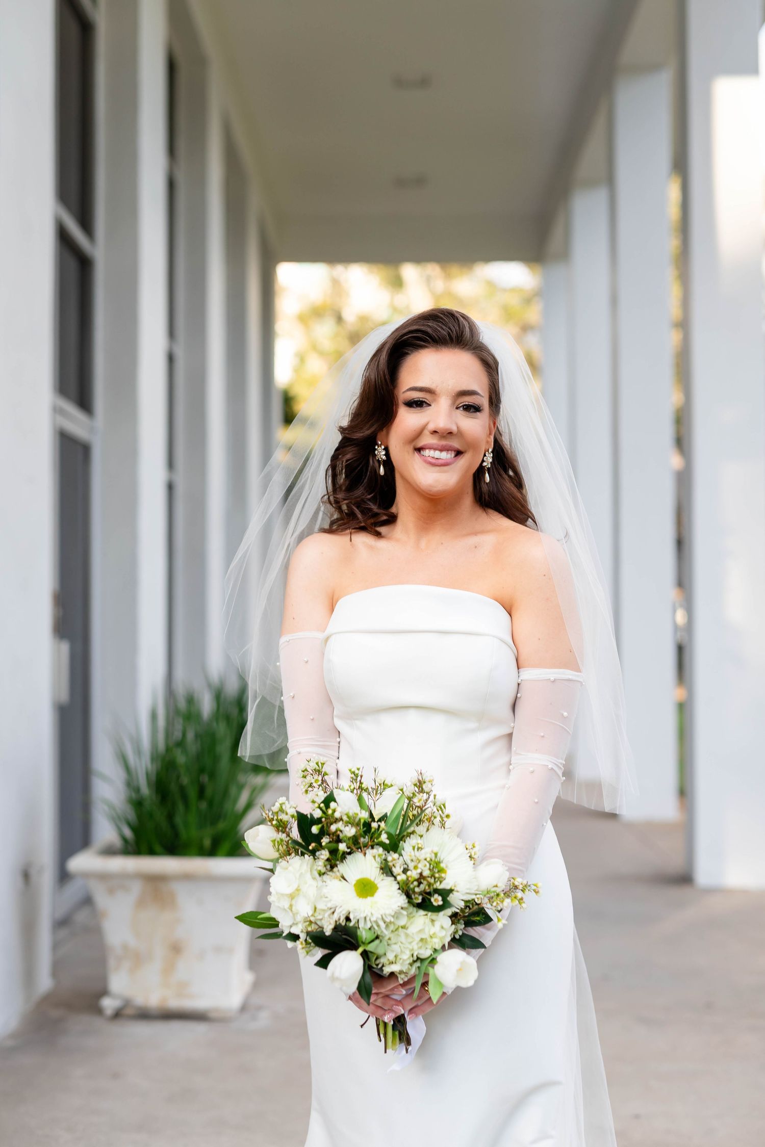 Elegant bride in strapless white gown and veil holds white floral bouquet while posing in a columned hallway on Boyshore Blvd in Tampa.