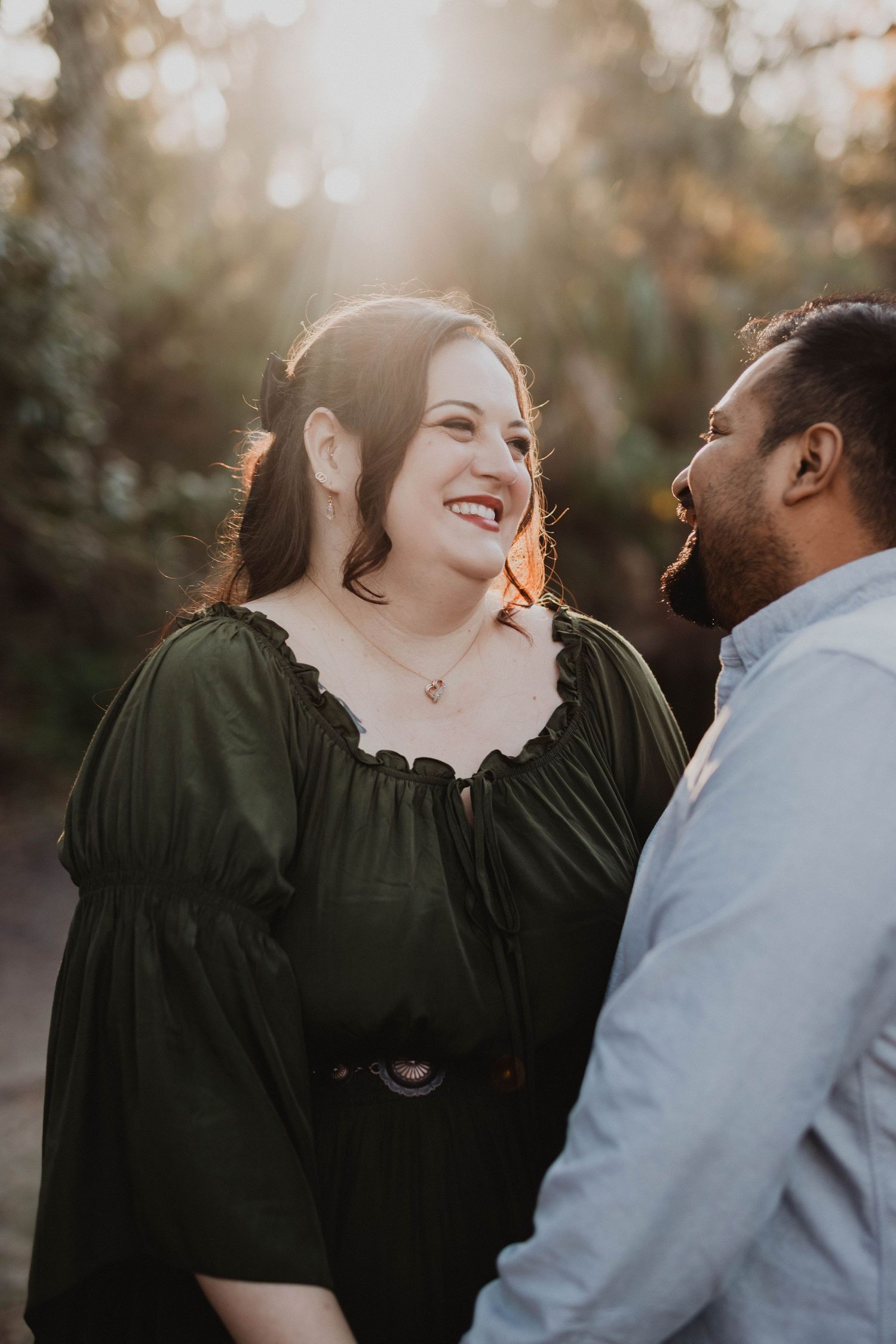 Couple laughing together outdoors in a sun-drenched forest during a golden hour engagement session.