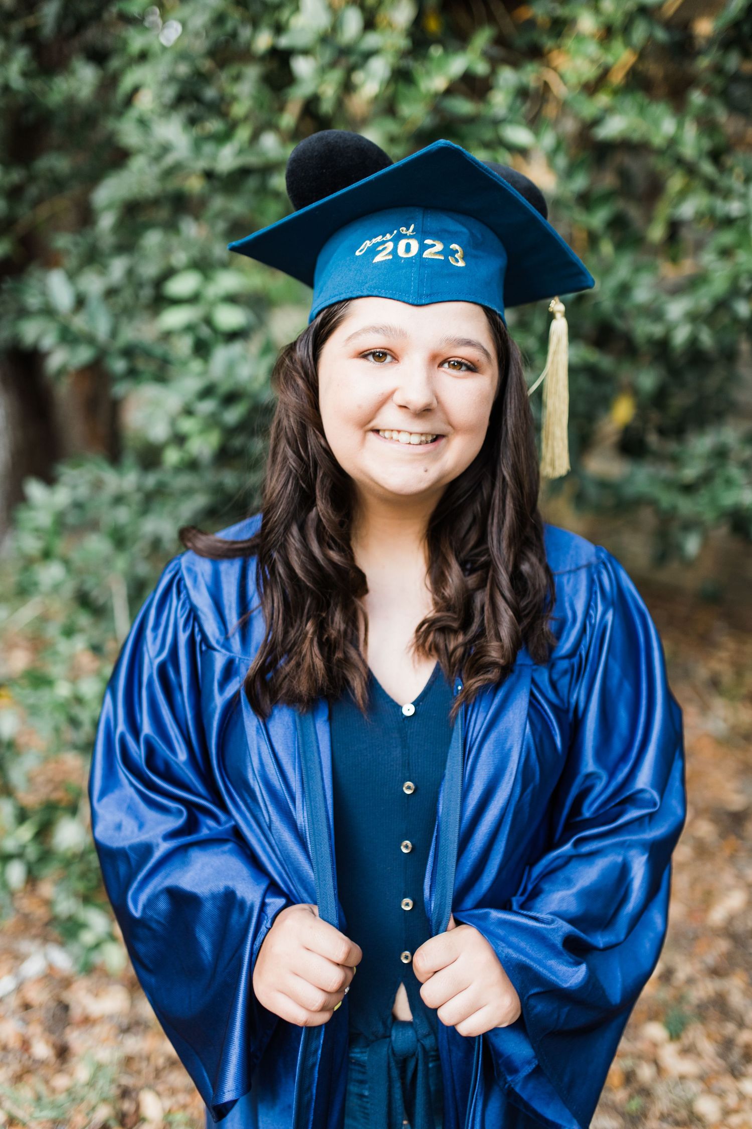 Smiling young woman in blue cap and gown celebrating 2023 graduation outdoors.