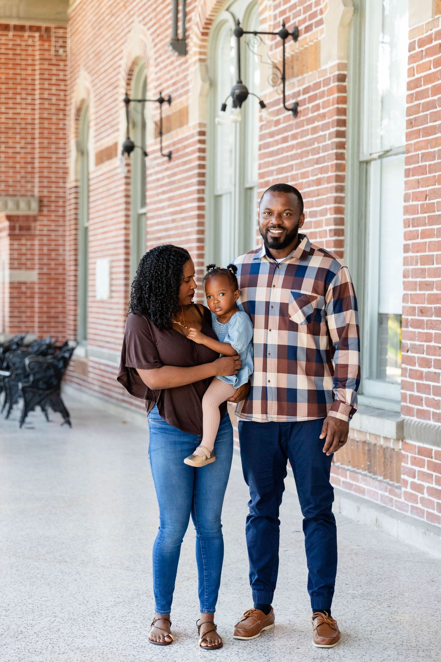 Smiling Black family with toddler poses in covered brick walkway, dad in plaid shirt at the University of Tampa.