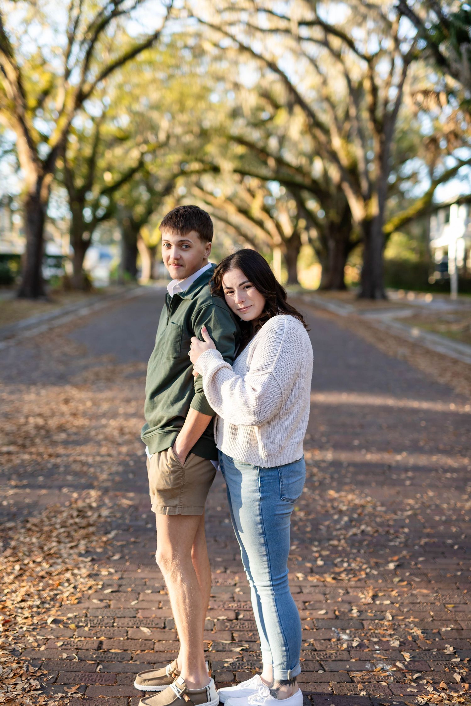 Couple posing together on a brick path under a canopy of oak trees in warm autumn light.